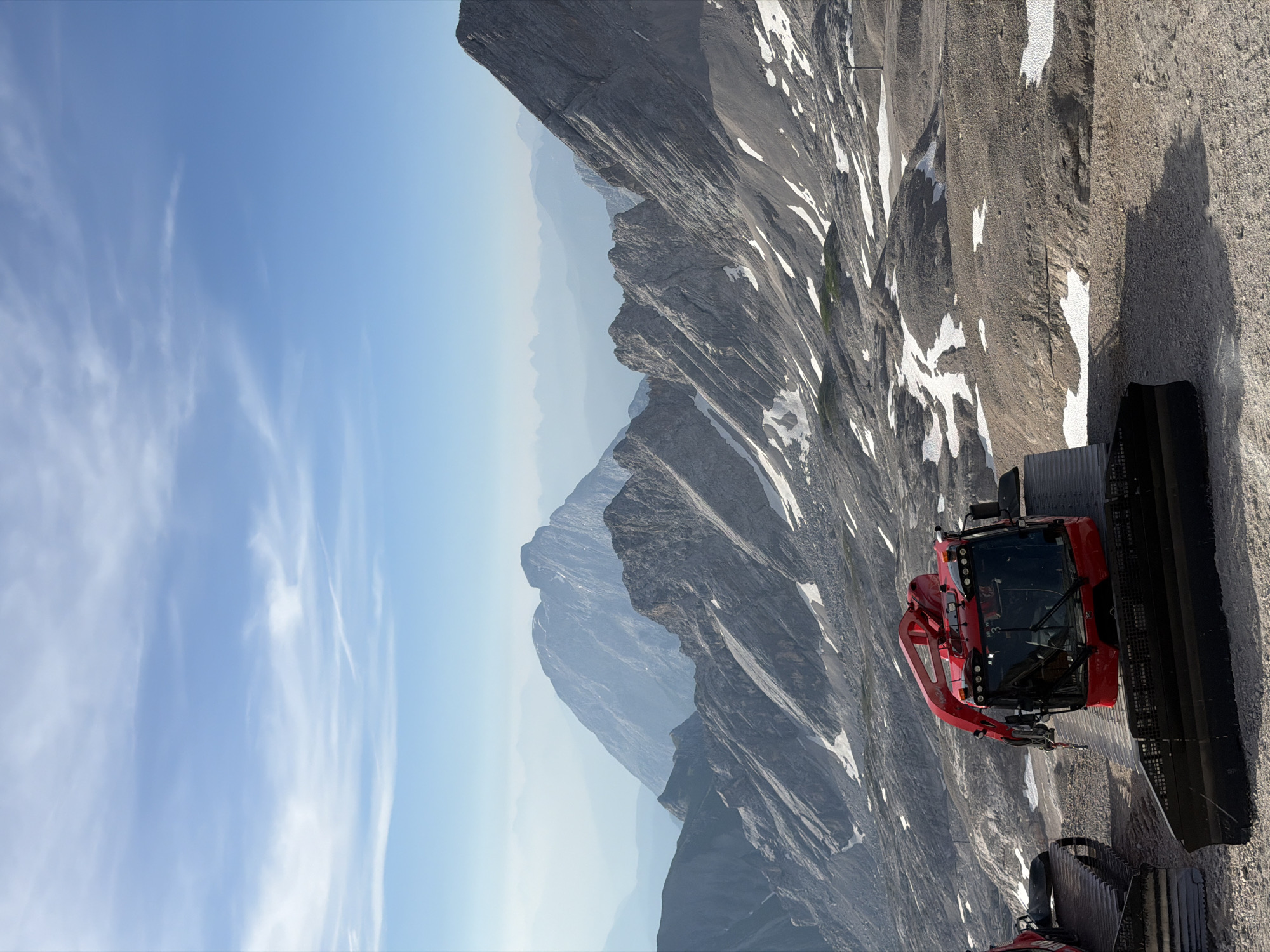This image captures a scenic mountain landscape with a red snow groomer in the foreground. The snow groomer is positioned on a gravelly surface, with its tracks visible. Behind it, rugged, rocky mountains rise, some with patches of snow, under a bright blue sky with wispy clouds. The scene suggests a high-altitude location, likely within the Bavarian Alps near Garmisch-Partenkirchen, Germany, possibly during the warmer months when snow may remain in shaded areas. There are no people visible in the frame. The background features layers of progressively hazy mountains, indicating a vast, expansive vista.