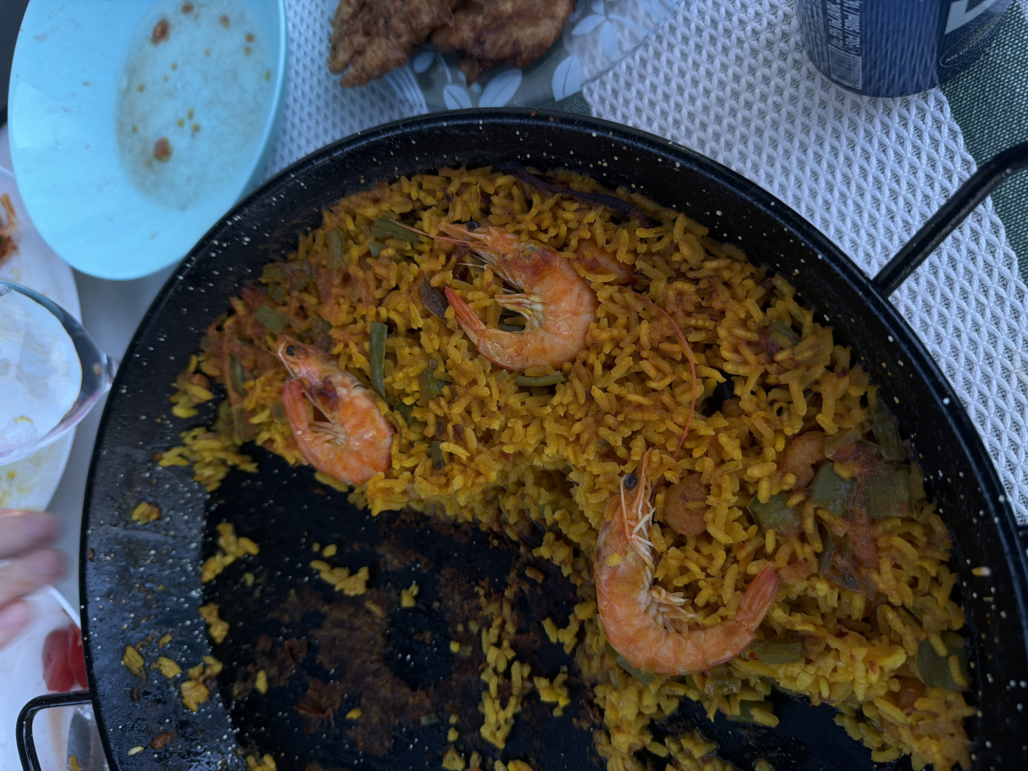 A close-up overhead shot shows a paella pan containing rice, shrimp, and green beans. The pan is half-empty, indicating that some of the food has been consumed. To the right of the paella pan, a blue bowl contains some residual liquid and food scraps. Further to the right, a plate holds a portion of fried food. The scene is set on a table with a textured white tablecloth, and a portion of a blue can with text is visible in the bottom right corner. In the background, a hand is reaching for food from a white plate, and a glass of water is also visible. The overall impression is of a casual meal being enjoyed outdoors, possibly at a restaurant or picnic.