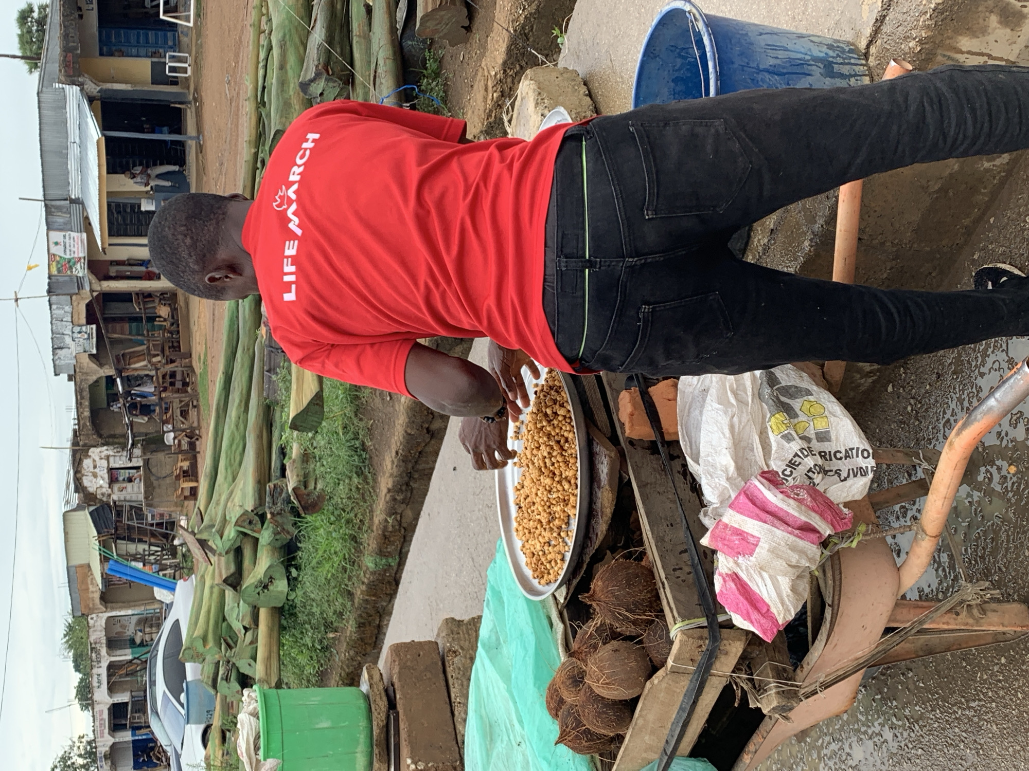 In Rafin Par, Nigeria, a man wearing a red shirt with the words "LIFE MARCH" in white text is standing outdoors next to a wheelbarrow. He is holding a silver platter filled with fried nuts. The wheelbarrow contains coconuts, a white bag printed with black and yellow text, and a pink and white striped plastic bag. A blue bucket is nearby. In the background are green logs and the storefronts of buildings across the street. The street surface appears to be wet.