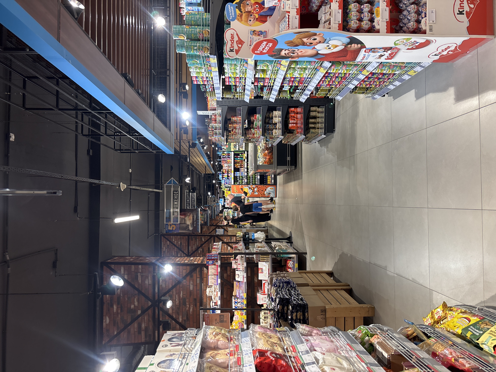 This is a photograph taken inside a brightly lit supermarket in Minsk, Belarus. The camera is positioned looking down an aisle that displays shelves of groceries. On the left, racks of packaged snacks and other food items are visible. To the right, shelves are stocked with canned goods and other merchandise, with a prominent display for Kinder products towards the front. In the mid-ground, a few shoppers are seen walking and browsing the aisles. The floor is tiled with large, light-colored squares, and the ceiling features exposed metal beams and lighting fixtures. The overall impression is that of a typical, well-stocked retail environment.