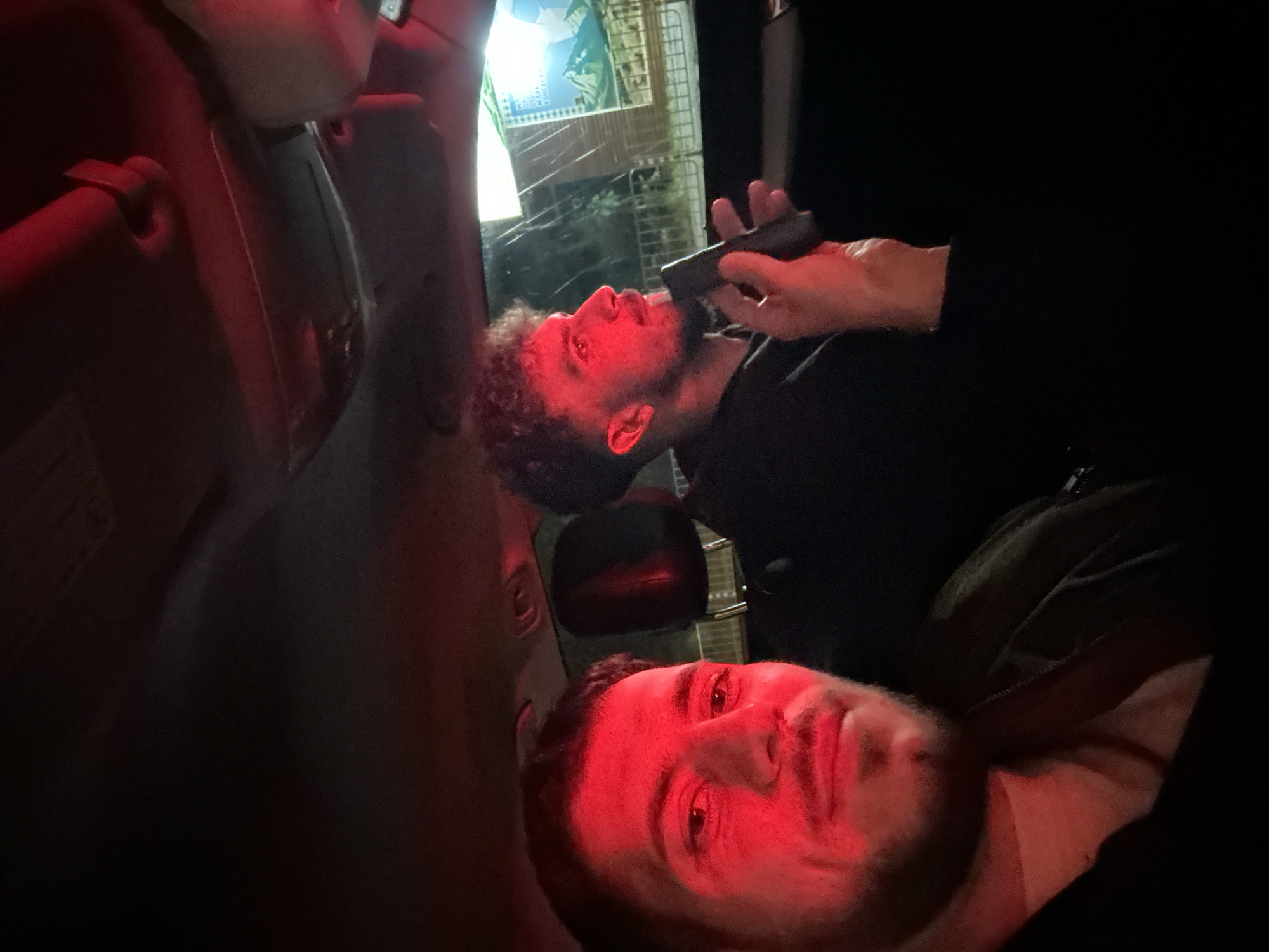 The image depicts two men inside a vehicle, potentially a car, at night. One man is lying down facing the camera, while the other man is visible with a hand holding an object, possibly a grooming tool, near his face. The scene is illuminated by red light, and in the background, there is an advertisement or poster behind the car window, hinting at a location in Sofia, Bulgaria.