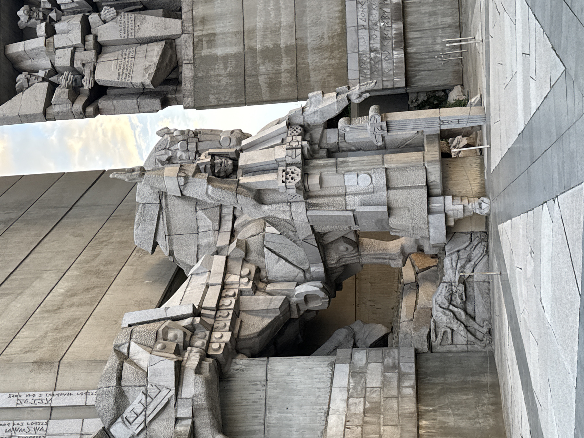 The image shows a close-up of a monumental sculpture or relief. The sculpture appears to be made of rough-hewn stone blocks. It depicts figures in action. The style of the sculpture is abstract or stylized. The sculpture is part of the Founders of the Bulgarian State Monument in Shumen, Bulgaria.