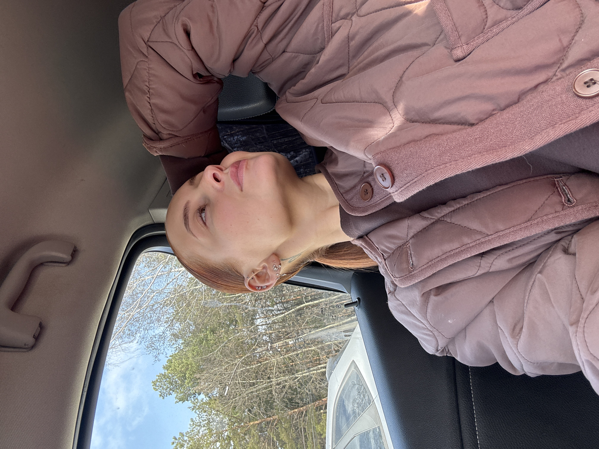 The image appears to be taken from inside a car. A woman with red hair is visible, wearing a brown quilted jacket. Outside the car window, a view of bare trees is seen under a blue sky. Based on the presence of Russian text on a sticker on the car window, it is probable that the location is in Russia. The absence of snow suggests it is not deep winter, but a season with bare trees. Given that the user specified the city is Chaikovsky, Russia, it's assumed that the picture was taken somewhere in the city or its surroundings.