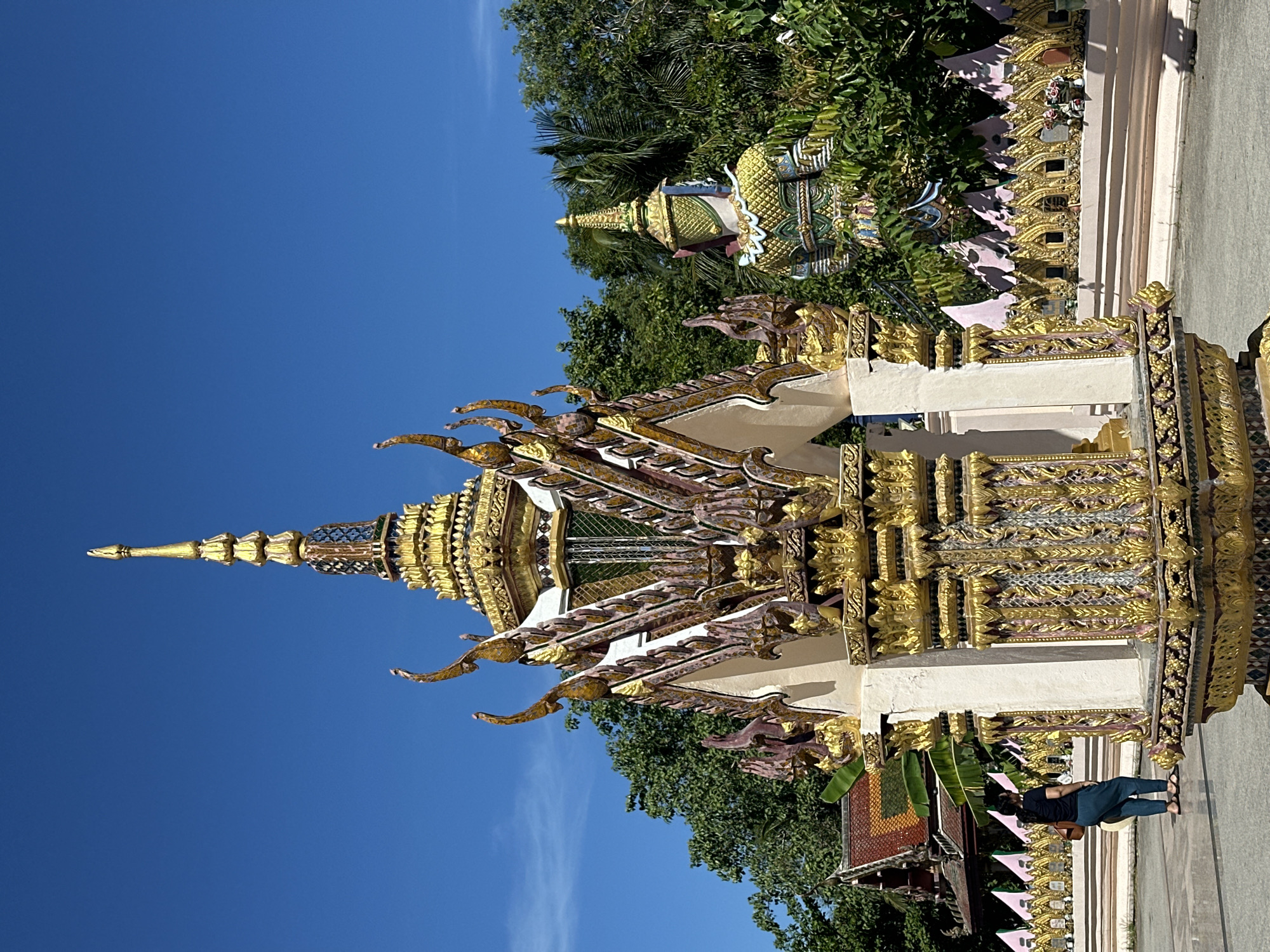 In Baan Plai Laem, Thailand, a woman stands on the paved ground of a temple complex, facing a decorative structure adorned with gold and intricate carvings. Behind her, a large, colorful statue of a guardian figure is partially obscured by lush green foliage. The temple grounds feature a decorative wall with many small, ornate structures. The sky is a clear, vibrant blue.