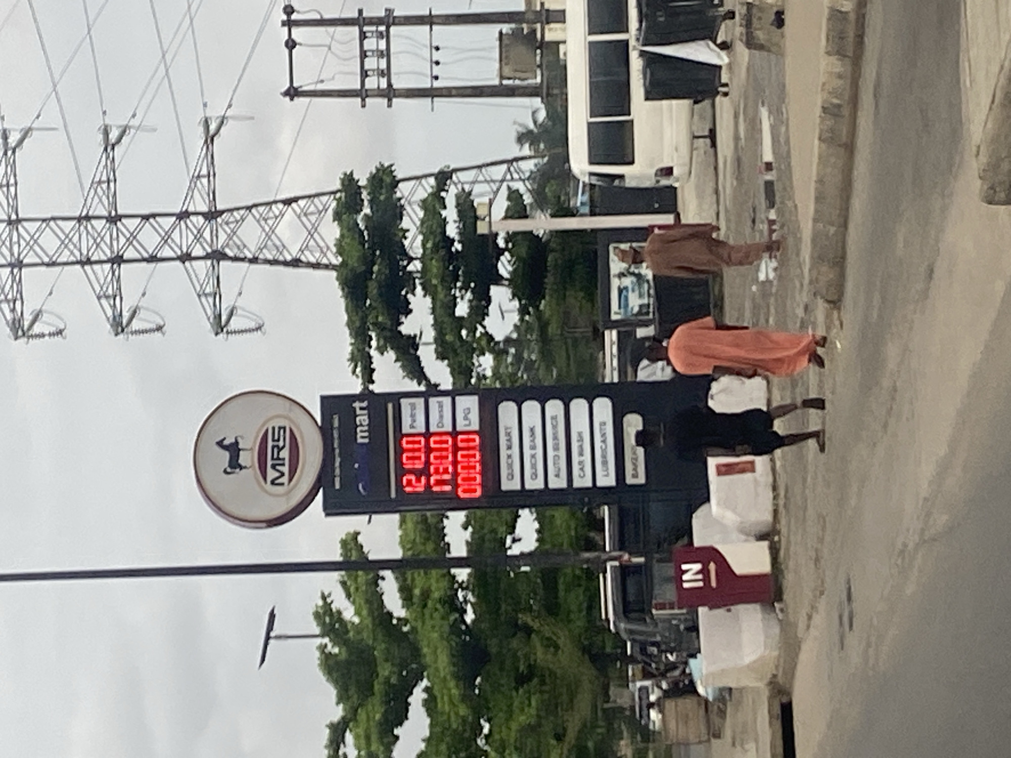 An exterior, medium shot captures a scene at an MRS gas station in Lagos, Nigeria. The gas price display board, a prominent feature, shows prices for Petrol, Diesel, and LPG, with the prices listed as 1210.0, 1730.0, and 0000.0 respectively. Below these prices, various services are listed: Quick Mart, Quick Bank, Auto Service, Car Wash, Lubricants, and Bakery. A tall electricity pylon with multiple crossbars and insulators stands in the background, suggesting a location near a power transmission line. A white minibus is parked to the right of the frame, with black bins and other assorted objects beside it. Two men are walking away from the viewer towards the right, one wearing an orange garment and the other a brown traditional outfit. Another man in a black t-shirt and shorts is seen walking towards the left. A red and white sign indicating "IN" is positioned in the foreground. The sky is overcast, typical of Lagos weather.