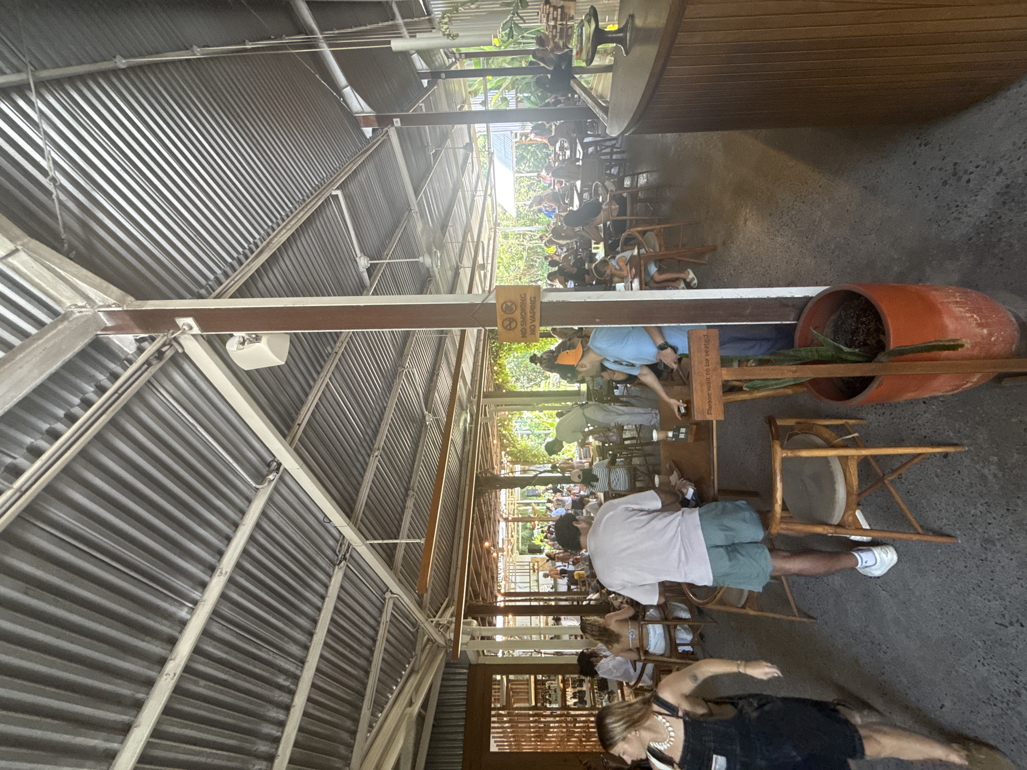 The image depicts an open-air restaurant in Kuta Selatan, Indonesia. The structure has a corrugated metal roof supported by wooden beams. Patrons are seated at tables and chairs throughout the space. A large terracotta pot sits near one edge of the image. People are present, some seated and others walking around. A "No Smoking/No Vaping" sign is visible.