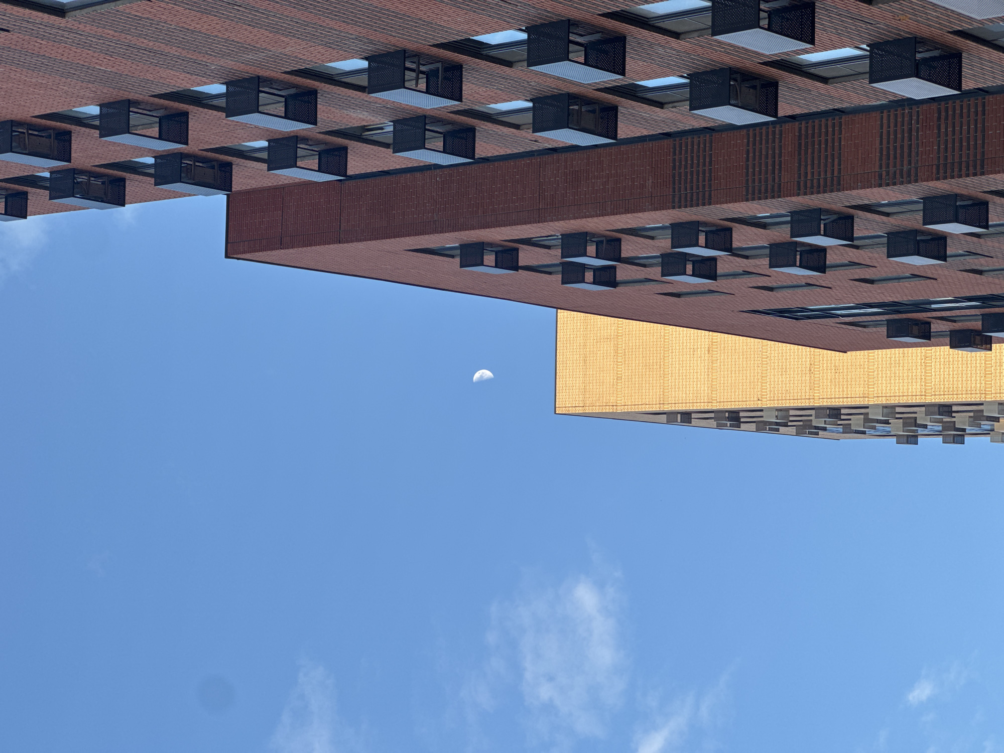 The image shows a view of buildings in Moscow, Russia. The perspective is angled upwards towards the sky. Two tall buildings are visible; one is made of brown brick with a grid of dark window frames, and the other is gold-colored and has a similar grid of window frames. A crescent moon appears between the two buildings against the blue sky with sparse clouds.