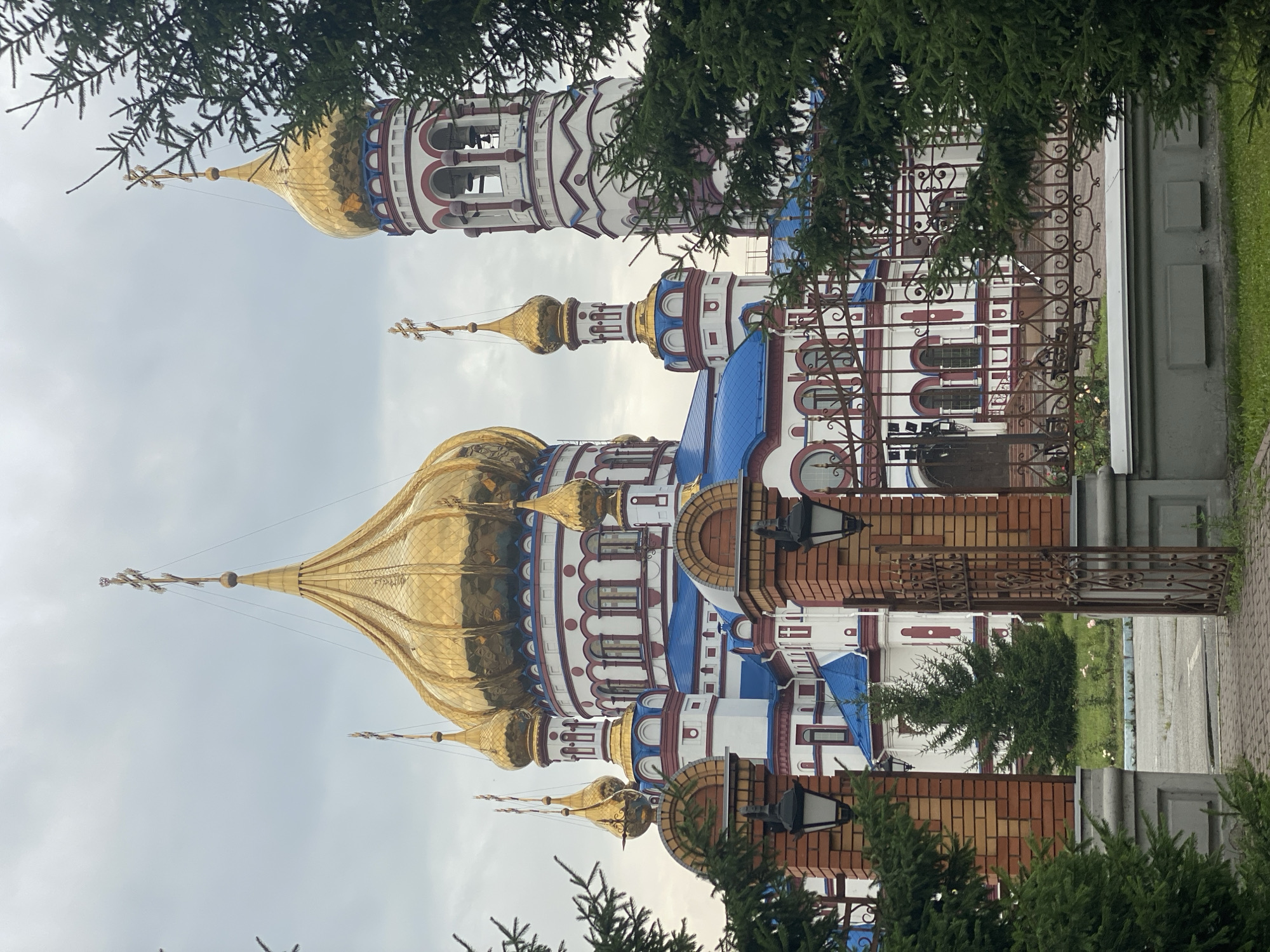 This is a photograph of the Cathedral of the Transfiguration in Novokuznetsk, Russia. The main subject is the cathedral itself, a large, ornate religious building with multiple golden domes topped with crosses. The cathedral's exterior is primarily white with red trim and blue roofs. To the right of the main building is a tall bell tower, also adorned with red trim and a golden dome. The foreground of the image features a red brick gate with an open metal gate, and several evergreen trees partially obscure the view of the cathedral. The sky is overcast.