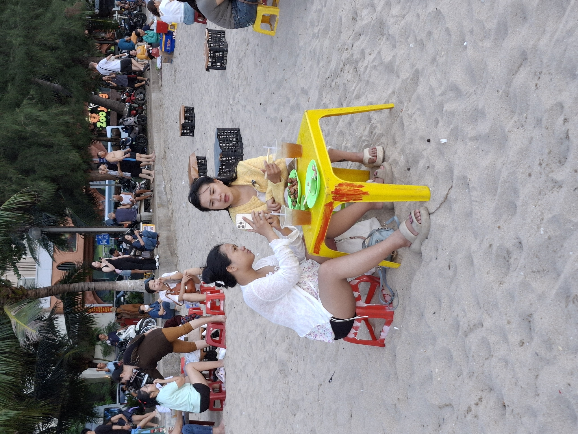 A group of people are gathered on a sandy beach. In the foreground, two women are seated at a bright yellow plastic table on the sand. The woman on the left is wearing a white sheer cardigan over a floral top and black shorts. She is holding a smartphone and looking at it. The woman on the right is wearing a yellow cardigan and appears to be eating. She holds chopsticks in her right hand. A plate of food and two drinks are on the table. Both women are wearing sandals. Red plastic stools are used as seating. In the background, other people are seen walking and sitting. Palm trees and other vegetation are visible, along with buildings and parked motorcycles. A sign with "Zoo" and a cartoon animal is lit up in the background. The scene appears to be a casual outdoor dining or gathering area on a beach, likely in Bac Nha Trang Ward, Vietnam, given the presence of palm trees, motorcycles, and the style of buildings and people.