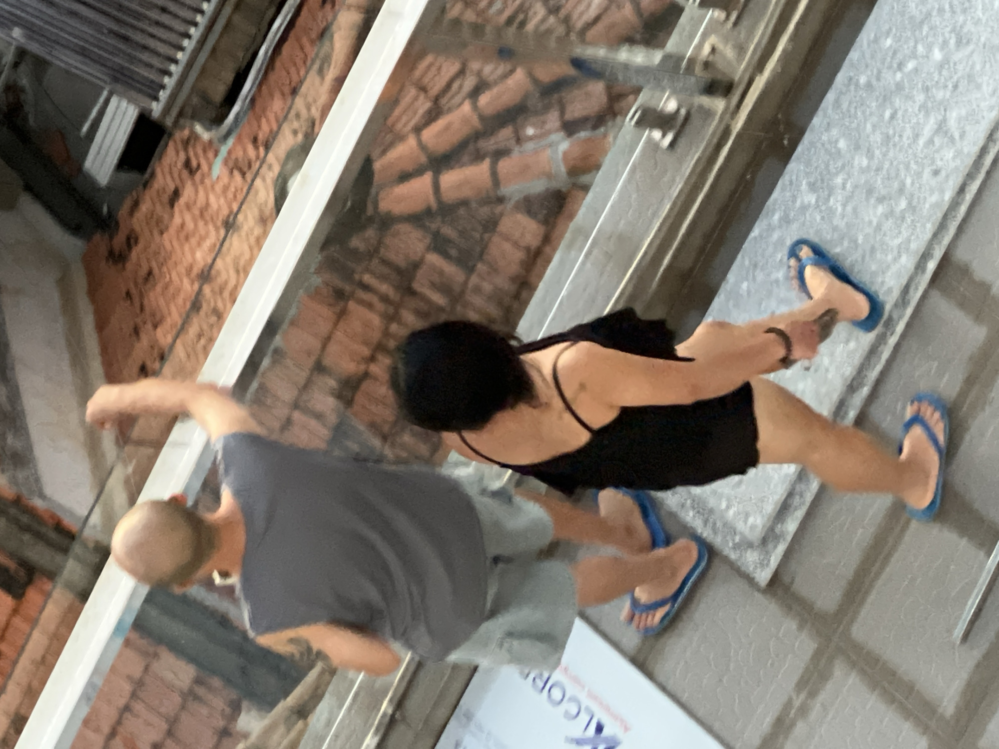 This image, taken in Nha Trang, Vietnam, shows two individuals walking down a staircase with a railing. The person in the foreground is a woman with dark hair wearing a black camisole and black shorts, with blue flip-flops. She is walking downwards, facing away from the camera. The person behind her is a man with a shaved head wearing a grey sleeveless t-shirt and light-colored shorts, also wearing blue flip-flops. He is also walking downwards and is slightly ahead of the woman. The staircase has a grey tiled surface. In the background, there is a view of terracotta roof tiles, indicating an outdoor setting, possibly in a more traditional architectural area of the city. A glass railing is visible between the two individuals.