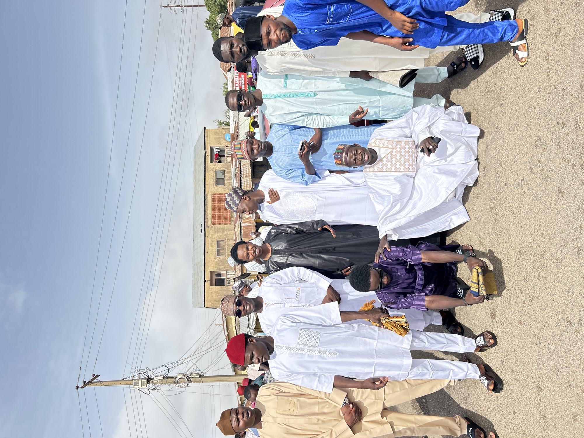 A group of approximately 12 men stands in a row on a paved road in Jos, Nigeria. The men are wearing various traditional African garments. Some wear hats and sunglasses. An urban setting with buildings and utility poles is visible in the background. The sky is lightly overcast.