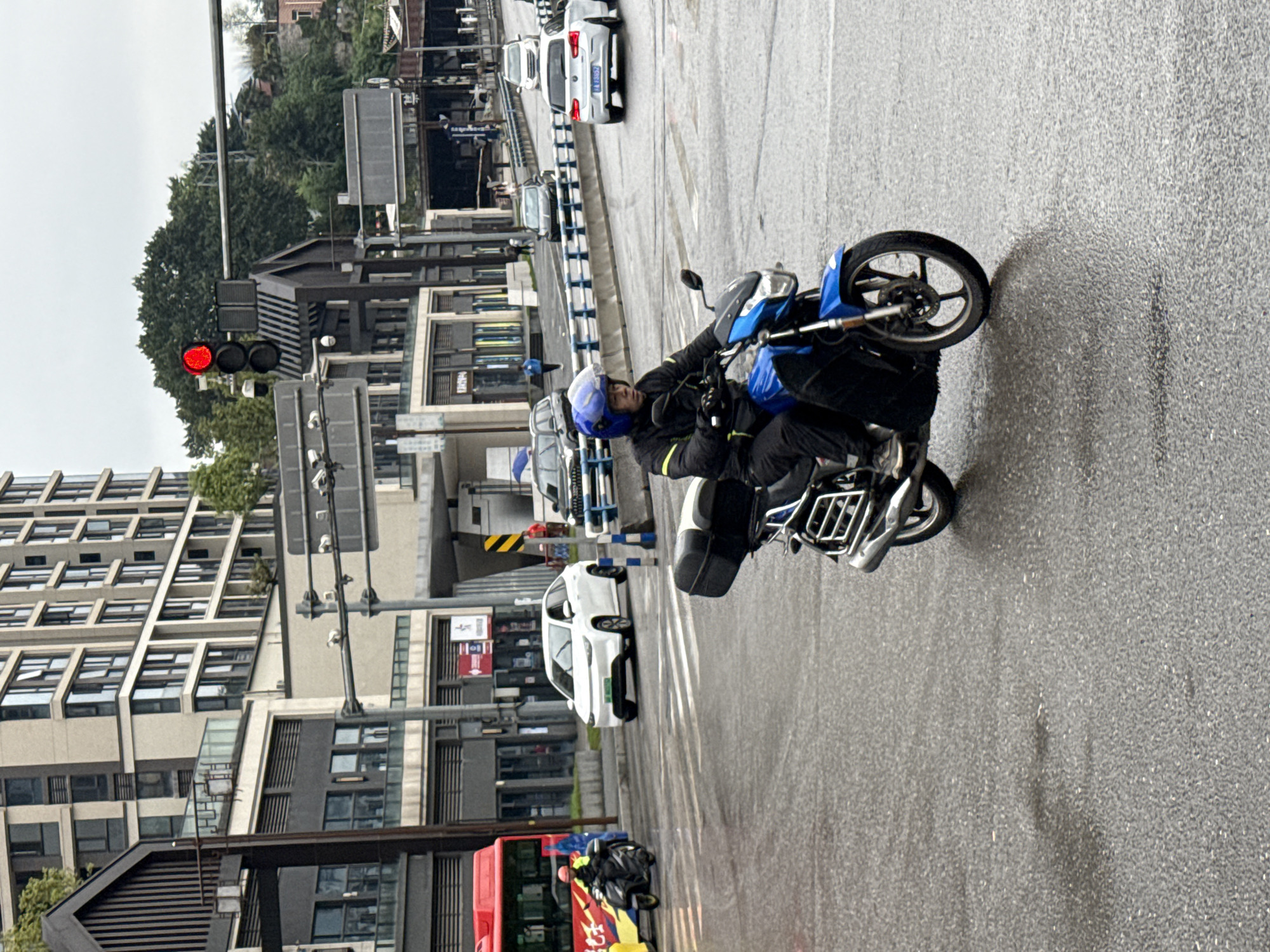 A man is riding a blue motorcycle on a wet road in what appears to be the Shapingba District, China. A red traffic light is visible, and there are buildings and vehicles along the street.