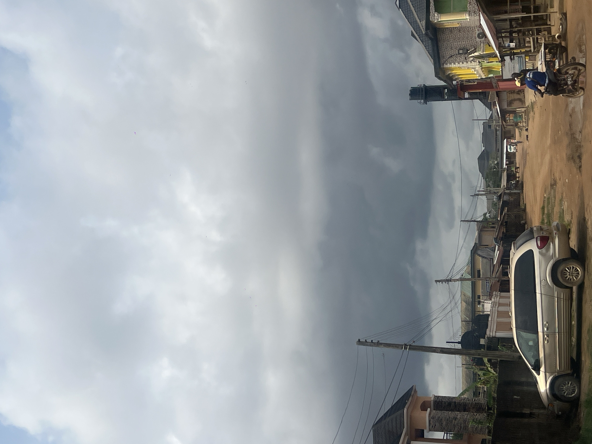 The image depicts a street scene in Egan, Nigeria. The sky is overcast with heavy clouds. On the right side of the frame, a tan minivan is parked on the side of the road. Buildings line the street, and utility poles with power lines are visible. Further down the street, a person is riding a motorcycle. The ground is dirt and appears slightly muddy.