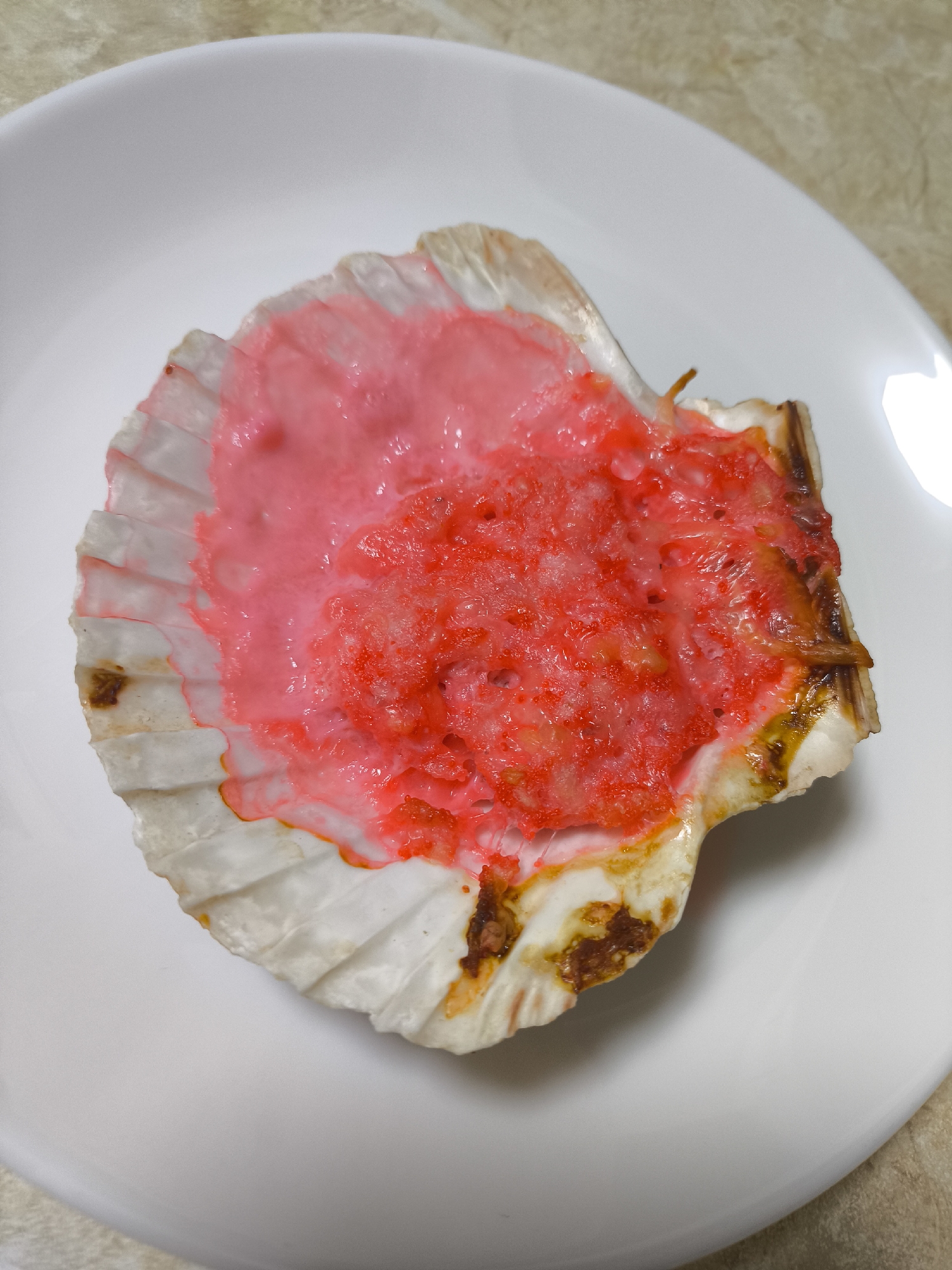A baked scallop, topped with a pink sauce and red granular topping, is presented on a white plate. The edges of the scallop shell appear browned and slightly burned. The background appears to be a plain countertop. The city of Khimki, Russia is not discernible in the image.