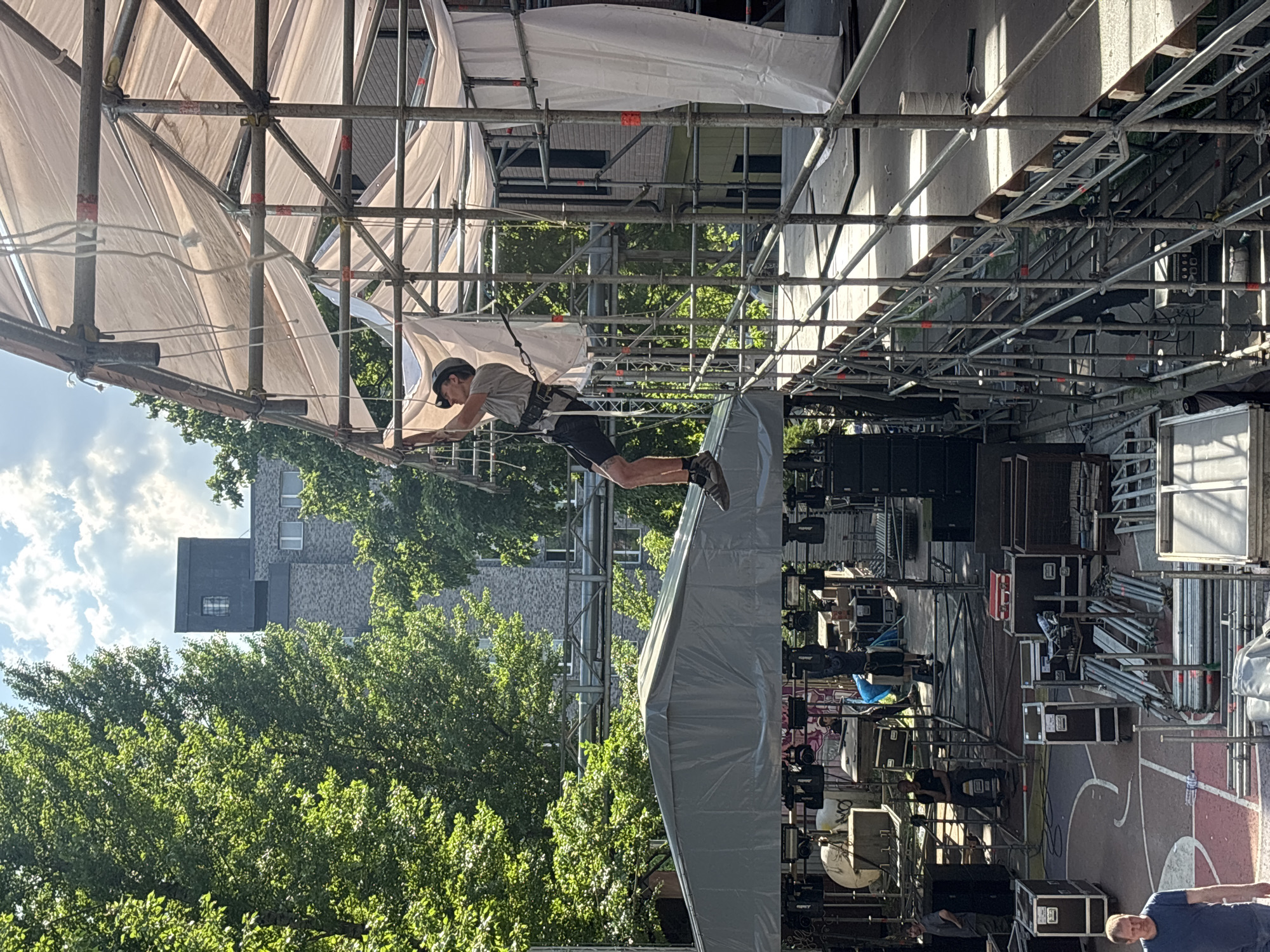 A man, wearing a gray shirt, black shorts, and a dark-colored helmet, is suspended from a scaffold by a harness, gripping the structure with his hands. The scaffold is covered with white fabric. Behind the scaffold are trees, a gray building, and a partly cloudy sky. The scene appears to be a setup for an event, with sound equipment, stacked crates, and additional scaffolding visible in the background. The location is Moscow, Russia.