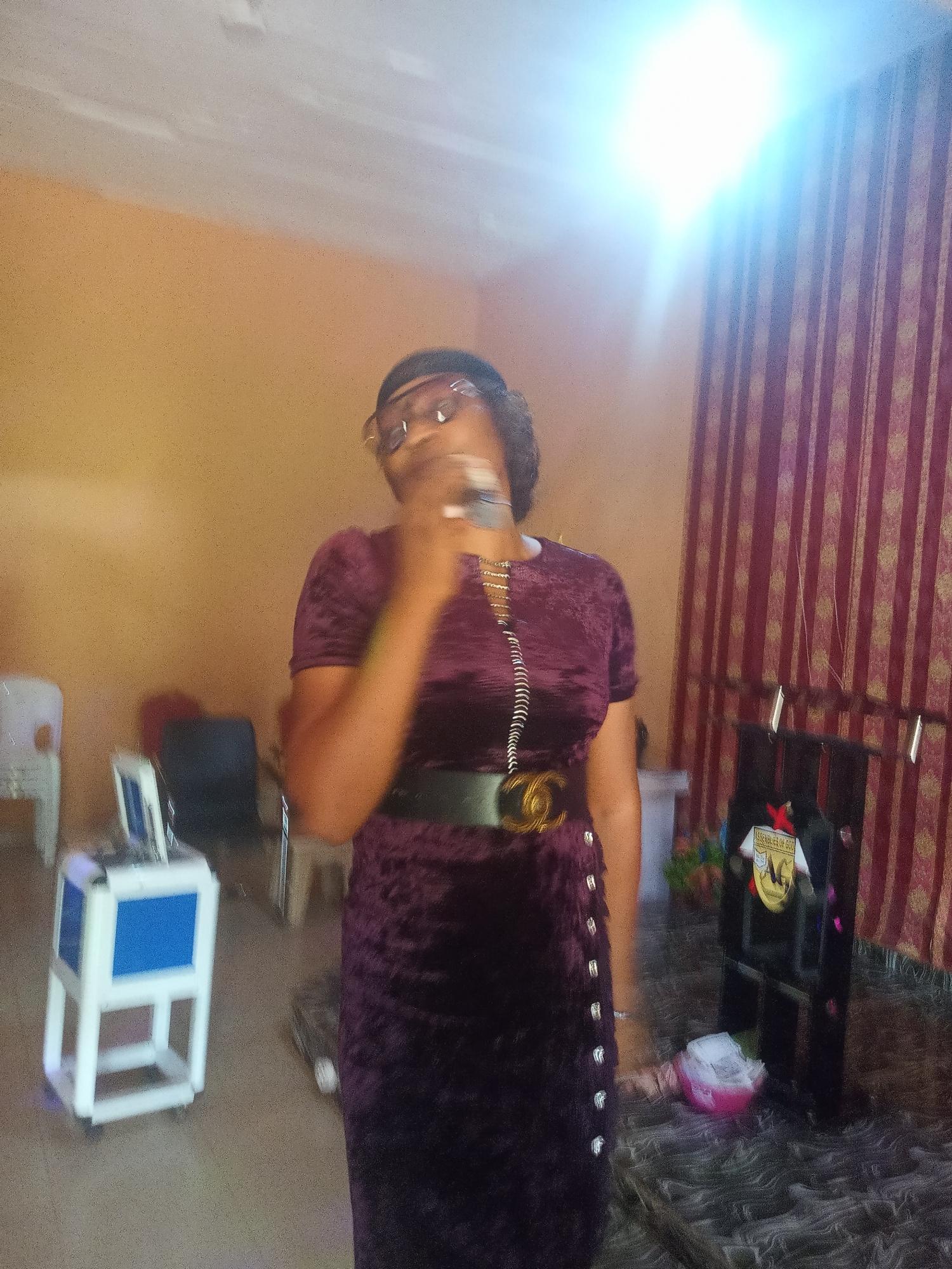 In what appears to be an indoor location, possibly within a church or meeting hall in Nkwelle Ezunaka, Nigeria, a woman is standing and holding a microphone. She is wearing a dark purple, possibly velvet, dress cinched at the waist with a wide, dark belt with a gold-colored buckle. She also wears glasses and a necklace. Behind her is a stand with the words "ASSEMBLIES OF GOD" on a logo or emblem. In the background, there are red and gold curtains, and to her left, there are pieces of equipment, possibly related to sound or audio-visual setup, including a white and blue structure. The room has orange walls. A bright light source is visible in the upper-right corner of the frame.