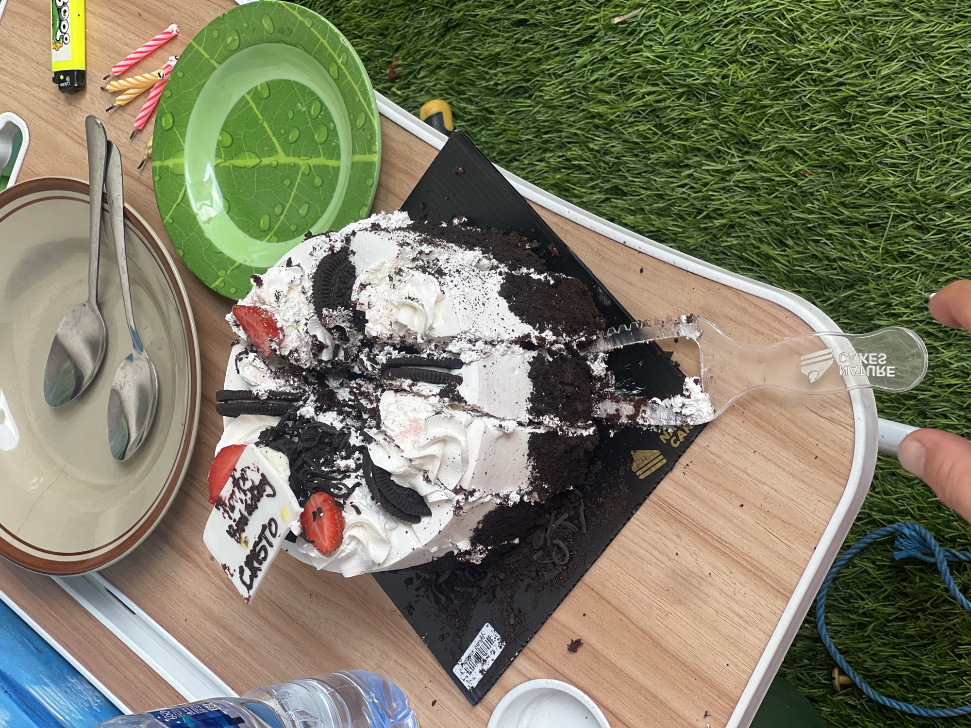 A partially eaten, chocolate and vanilla cake with strawberries and Oreos sits on a black cake board on a light-colored wooden table. A white decorative topper with "Happy Birthday CRISTO" written in black icing is placed on the cake. To the left of the cake is a clear plastic water bottle. To the right of the cake are two spoons on a brown-rimmed plate, a green plate with a leaf design, and several birthday candles. In the background, green grass and a yellow and black object are visible. The scene appears to be outdoors, possibly in Denpasar, Indonesia, given the context of a birthday celebration.