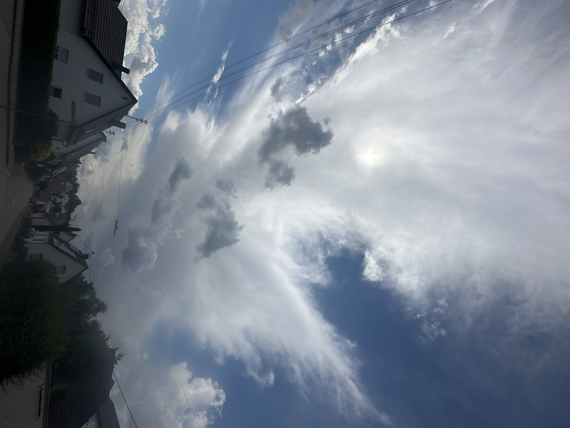 The image shows a suburban street in the city of Günzburg, Germany, under a dramatic sky. The perspective is looking up towards the sky from ground level, with houses and trees occupying the lower portion of the frame.
The sky is characterized by a striking mix of bright blue patches and a vast expanse of white and grey clouds. Wispy, feathery clouds stretch across the upper half of the sky, illuminated by sunlight that creates a hazy glow in the center. Towards the right and lower parts of the sky, darker, more voluminous clouds are visible, suggesting the possibility of impending rain or a storm. Power lines are strung across the sky, diagonally crossing from the left side.
In the foreground, several residential buildings with dark tiled roofs are visible. They are typical of European suburban architecture, with gabled roofs and visible windows. Some houses are bordered by neatly trimmed green hedges. A paved road or sidewalk runs through the center of the image, leading into the distance where more houses and trees can be seen, fading into the overcast sky. There are no people or vehicles clearly depicted in the scene. The overall impression is one of a dynamic and somewhat moody weather condition in a peaceful residential area.