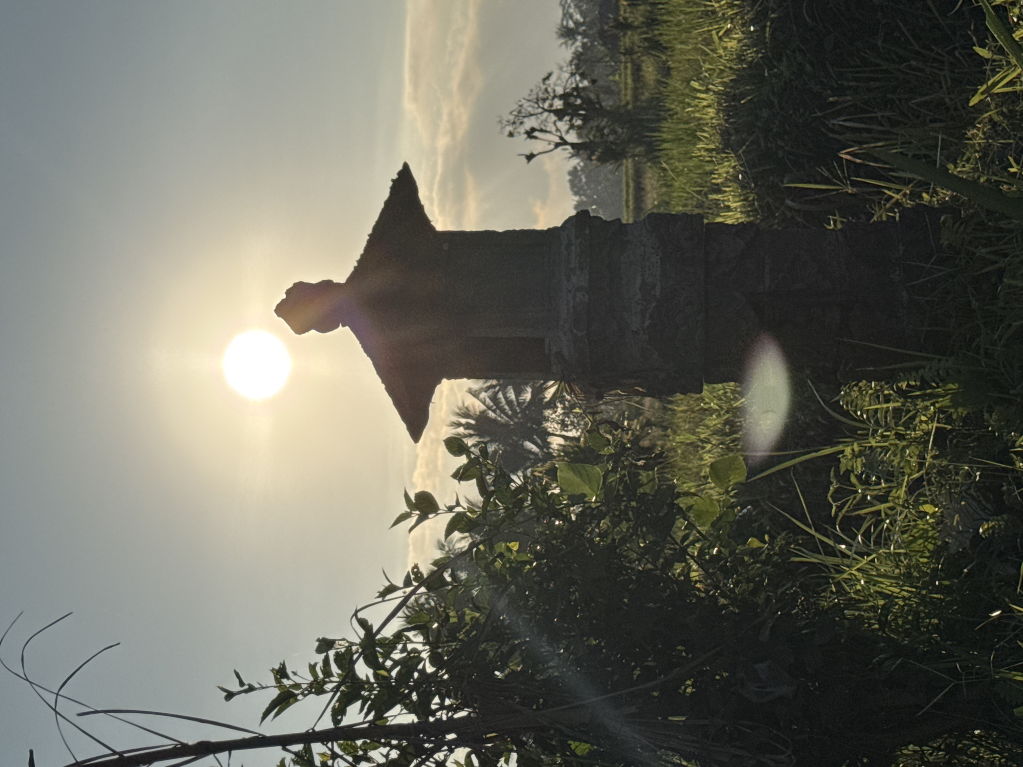 In Ubud District, Indonesia, at sunrise, a stone pagoda-style lantern stands silhouetted against the bright sun in a verdant field. Lush green grass and foliage surround the lantern, with hints of palm trees and distant structures visible in the background. The sky is a soft gradient of blue and yellow, with a few scattered clouds. Lens flares appear in the image due to the direct sunlight.