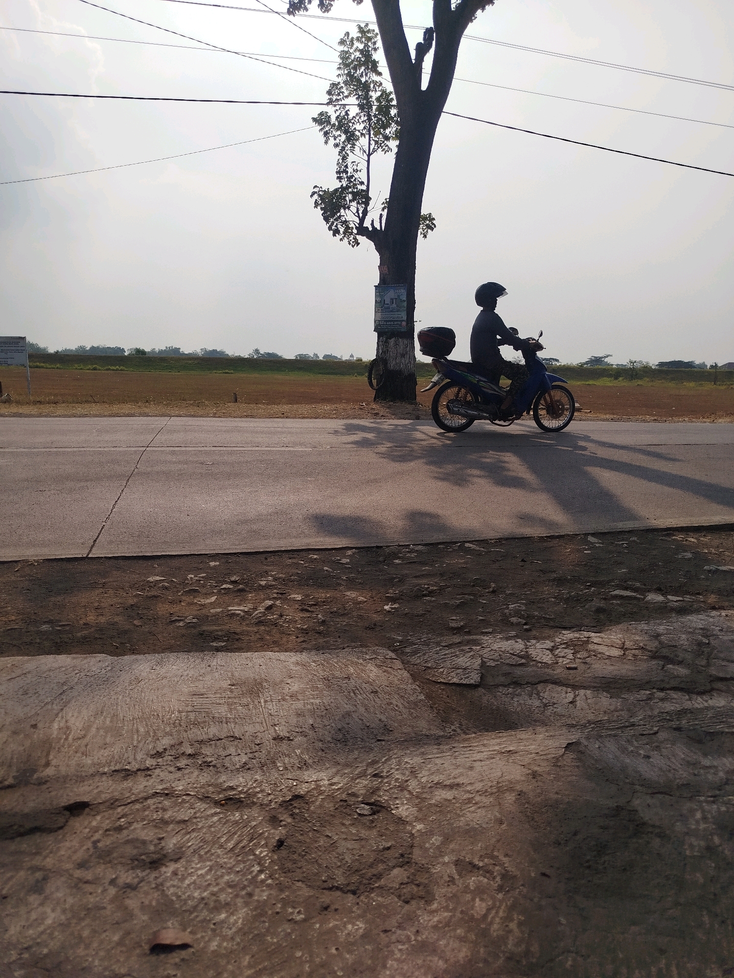 A lone rider on a blue and green motorcycle wearing a black helmet and dark clothing is captured in profile, moving from left to right across the frame on a paved road. The motorcycle has a black storage box on the rear. The scene is set in Ngreco, Indonesia, under a hazy, overcast sky. A large tree with sparse foliage stands to the left of the road, its shadow and the motorcycle's shadow stretching across the asphalt. The background features a flat, dry field and distant trees. The foreground is dominated by rough, textured ground with scattered rocks and debris, suggesting an unpaved shoulder. Power lines are visible crisscrossing the sky above the scene. A sign is affixed to the tree on the left.