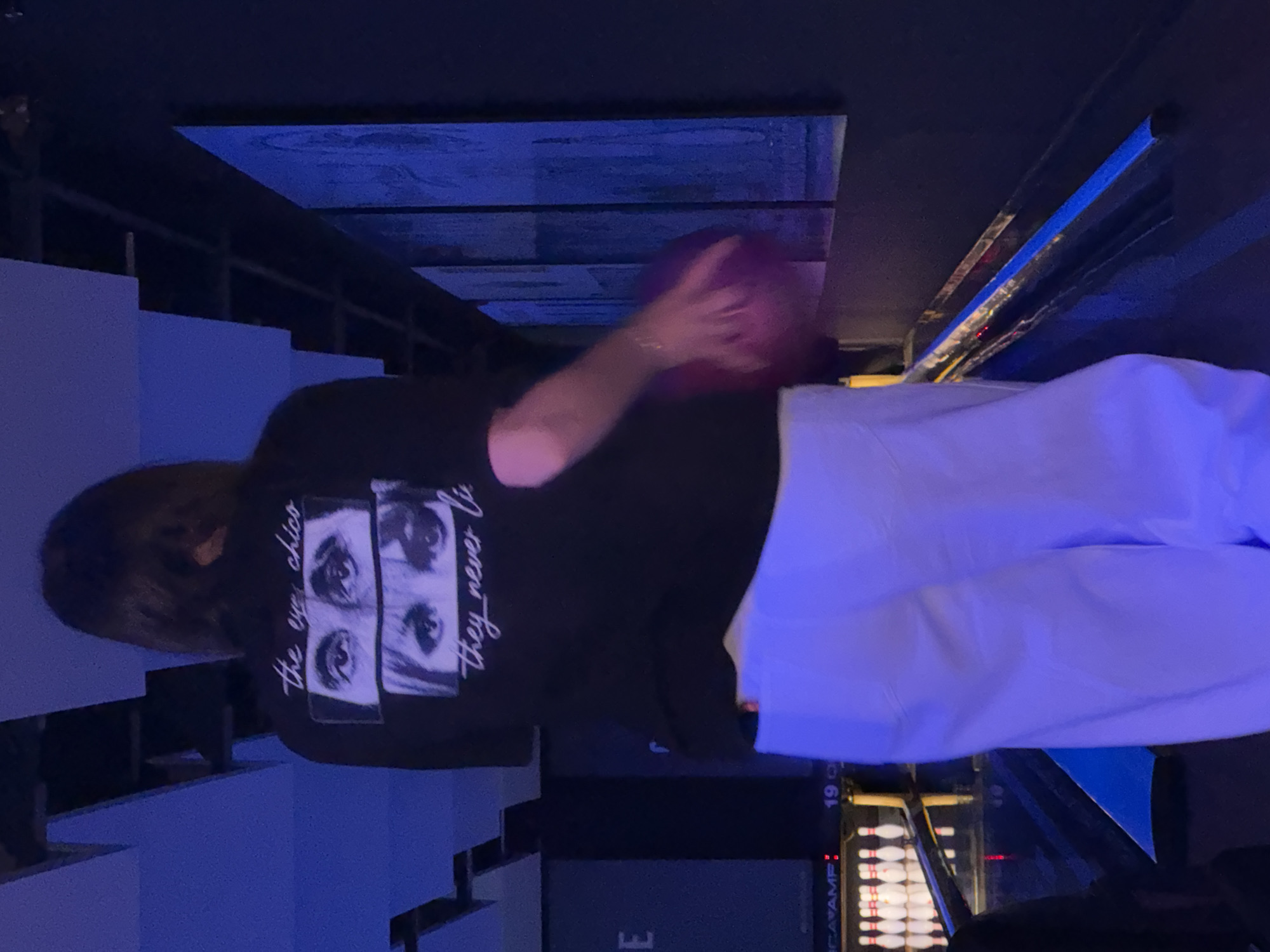 The image captures a person from behind at a bowling alley in Sofia, Bulgaria, illuminated by dim, ambient blue and purple lighting. The individual has dark hair and is wearing a black short-sleeved t-shirt with a graphic on the back depicting two pairs of eyes—one male's above a female's—and text that reads "the eyes chico" and "they never lie." They are also wearing white trousers. The person holds a purple bowling ball in their right hand, positioned as if preparing to bowl. In the background to the left, a set of white bowling pins stands arranged at the end of a lane. Above the pins, a digital display shows "19 O". The immediate environment includes a bowling lane, a dark wall with vertically oriented blue-lit panels on the right, and a ceiling with dark, recessed panels.