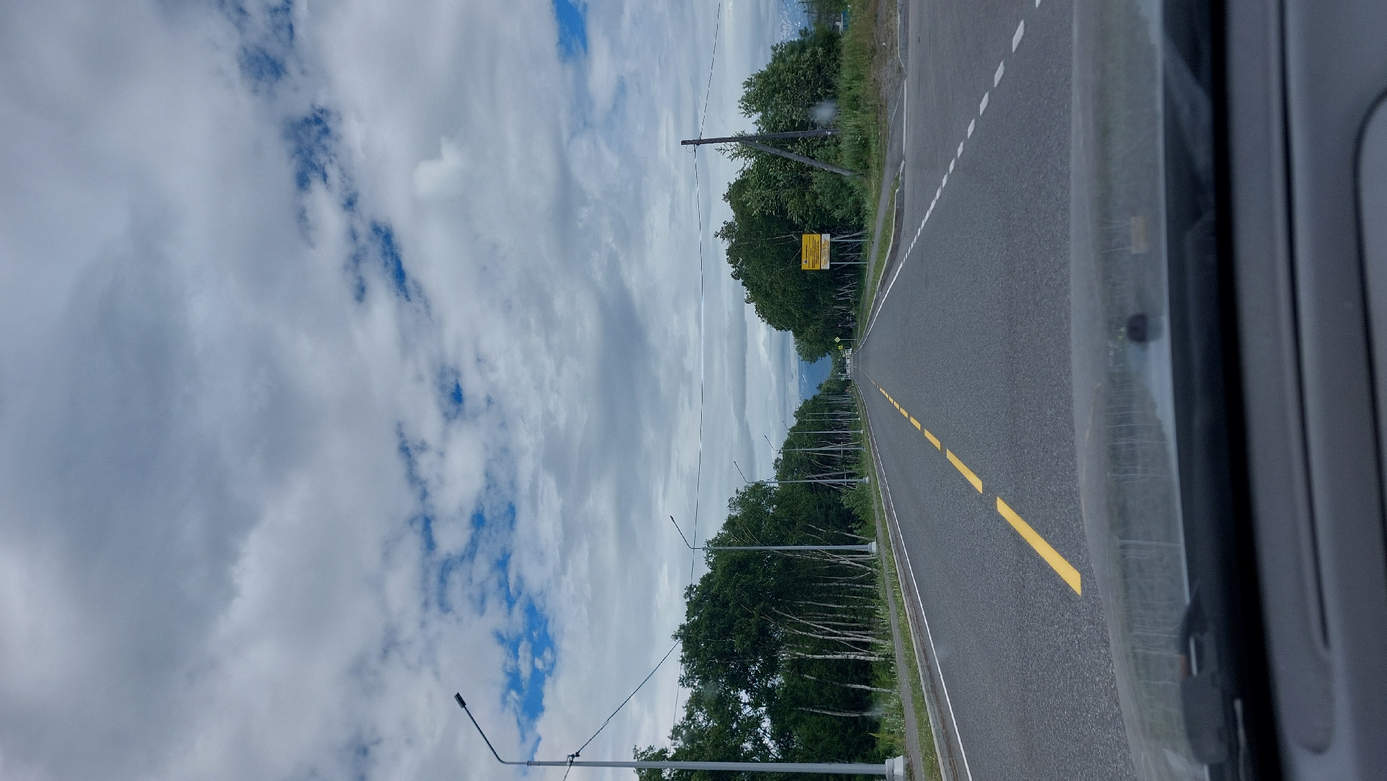 This is a vertical, outdoor, daytime, eye-level shot of a paved two-lane road in Koryaki, Russia. The road is bordered by trees on both sides, with a scattering of utility poles and streetlights. The sky is overcast with scattered blue patches. The foreground is dominated by the dashboard and windshield of a vehicle, implying the photo was taken from inside a car. The road extends into the distance, appearing to lead towards a town or more populated area. A yellow broken line marks the center of the road. A yellow sign is visible on the right side of the road, but its text is illegible. The overall scene suggests a journey through a rural or semi-rural area.