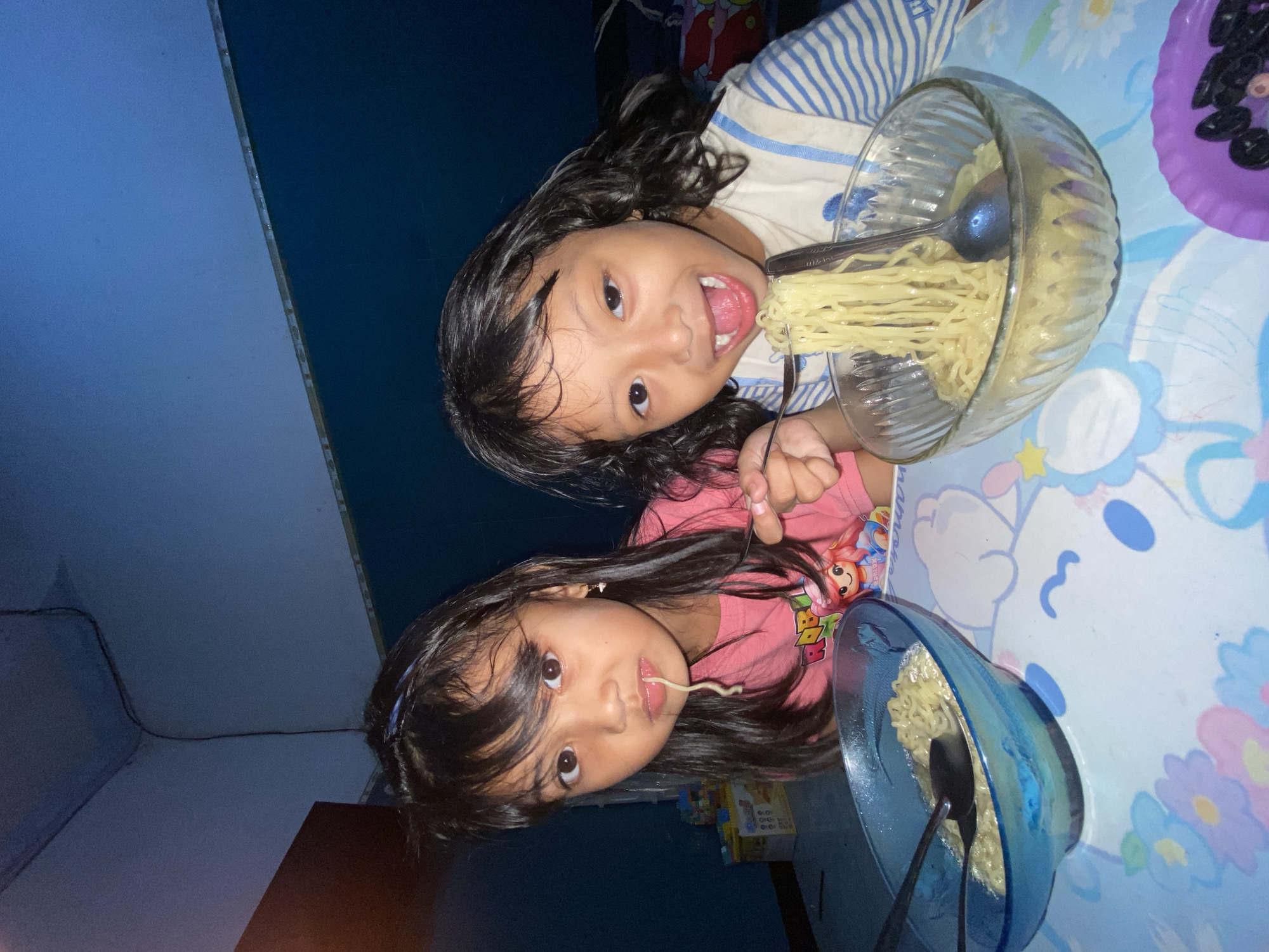 Two young girls are indoors, photographed from a low angle. The top girl, wearing a blue and white striped shirt, faces the camera with an open mouth, holding a fork with noodles. A glass bowl full of noodles sits in front of her. The bottom girl, wearing a pink t-shirt, eats a single noodle with a spoon in a blue bowl. They are at a table with a light blue tablecloth decorated with flowers and cartoon characters. There is also a purple dish with dark objects near the top girl. The background shows a dark wall and ceiling. Based on the location details, it is taken in Gelam, Indonesia.
