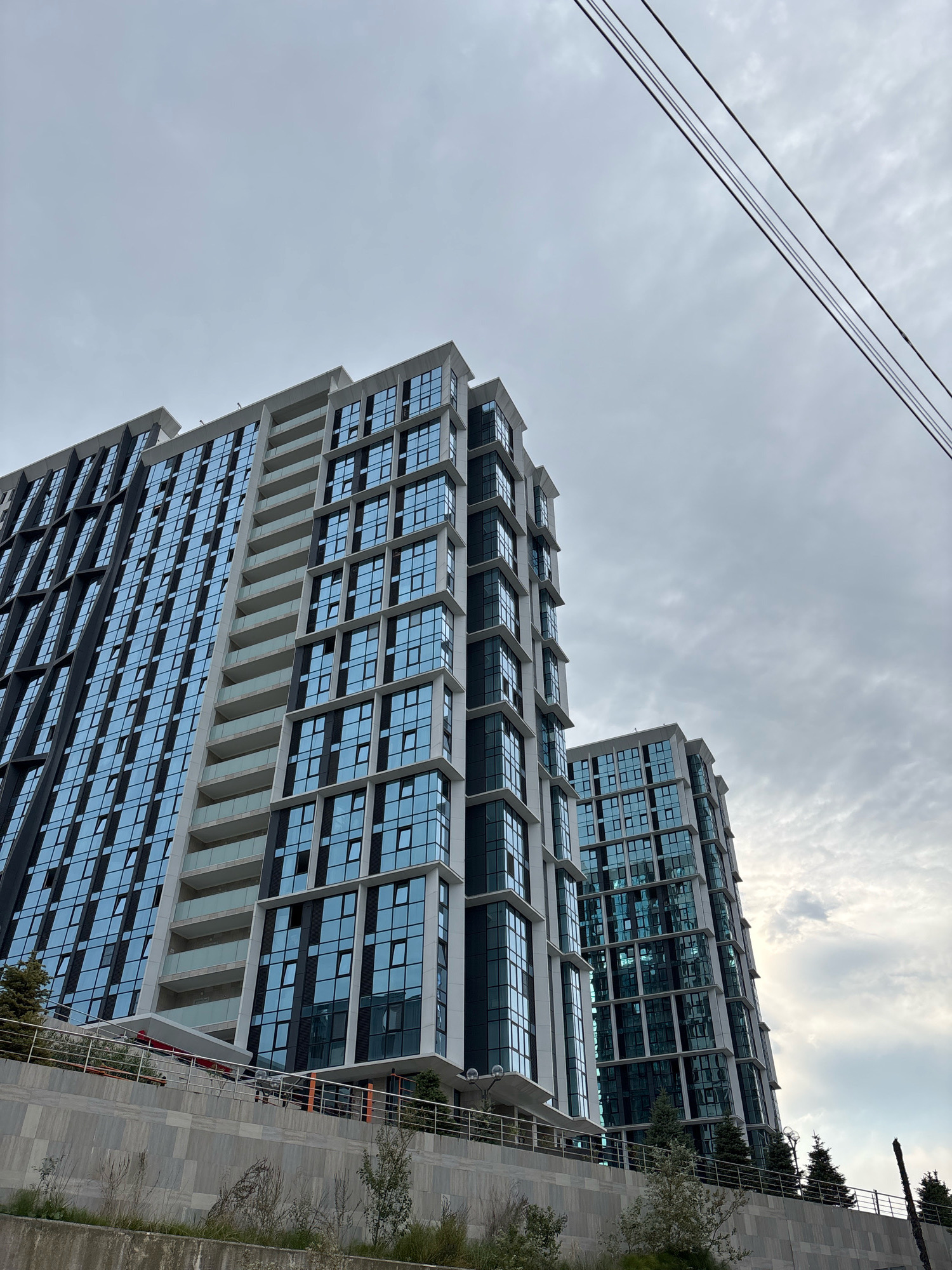 The image shows a low-angle view of two modern, multi-story apartment buildings with glass facades. The buildings have a geometric design with balconies and window grids. Overhead power lines are visible against a cloudy sky. In the foreground, there is a retaining wall with some vegetation and a metal railing. The scene appears to be in an urban or suburban environment. The buildings are modern residential structures. The location is identified as Sochi, Russia. There are no people or visible actions in the image.