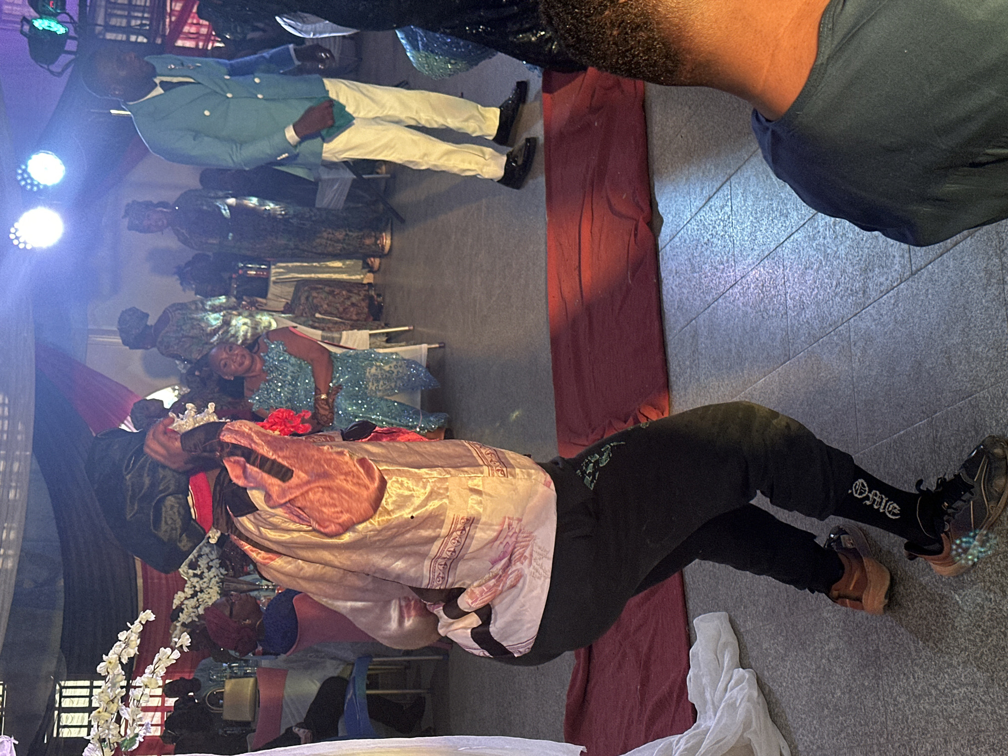 An indoor scene captures a vibrant social gathering, possibly a celebration or cultural event. In the foreground, a person with dark skin is prominently featured, wearing a light pink and gold patterned tunic over black pants and sneakers. They are holding a white floral arrangement near their head, which is adorned with a small crown or tiara. The text "OME" is visible on their black socks.
To the left and slightly behind, a woman in a shimmering blue, sequined dress smiles brightly towards the camera, conveying a sense of joy. Further back in the mid-ground, a man in a light blue blazer, white pants, and black dress shoes stands formally, holding a green piece of fabric, possibly a handkerchief. Other attendees, some dressed in patterned fabrics and headwraps, are visible in the background, suggesting a diverse and festive crowd.
The setting features a grey tiled floor, and the area is illuminated by multiple bright spotlights from the upper left, creating a lively atmosphere. Red and dark drapes are part of the event's decoration, along with various floral arrangements including white and red flowers. The attire and general ambiance suggest a celebratory and culturally rich event, possibly of West African origin. The time of day is not discernible due to the indoor lighting.