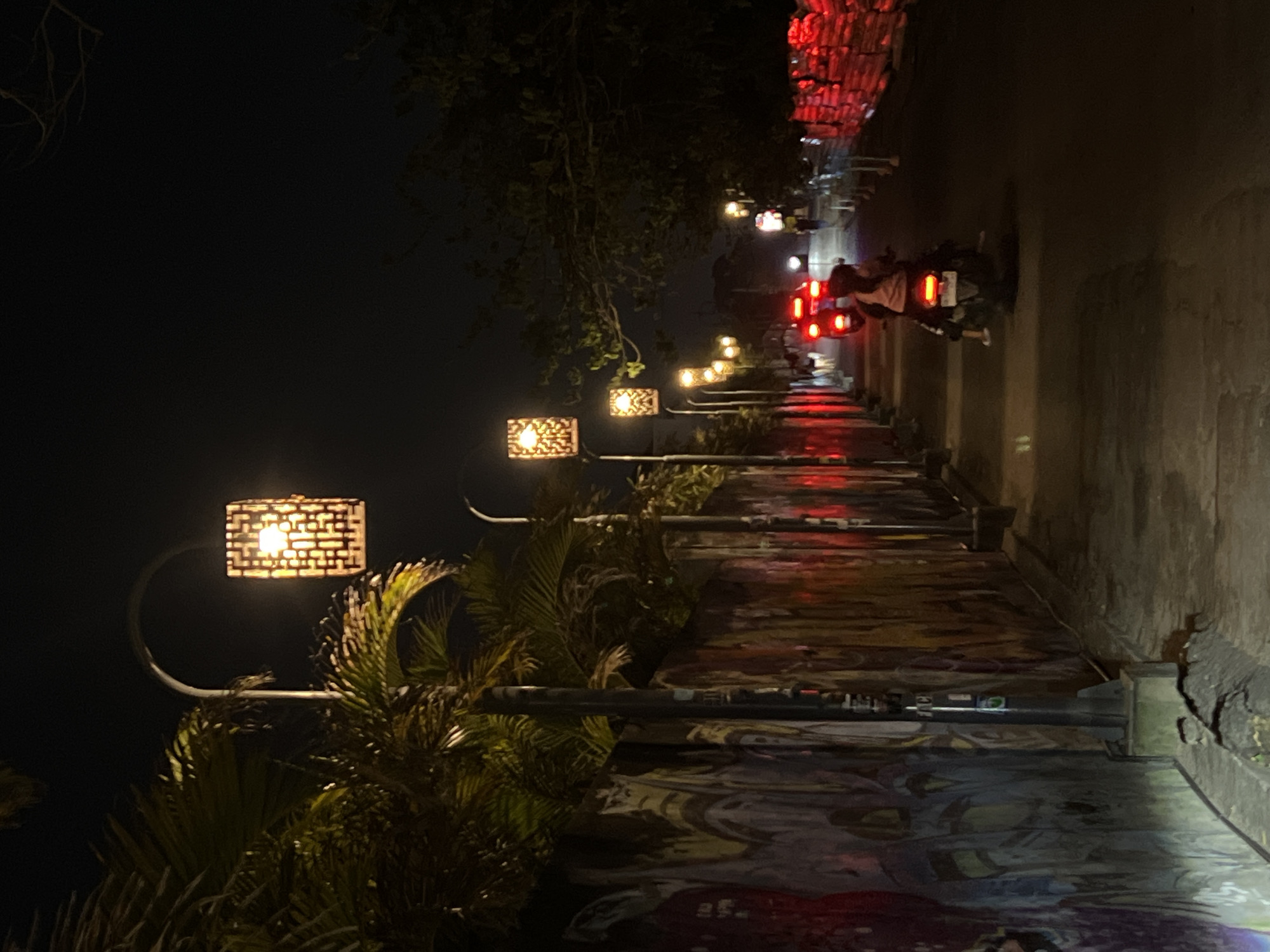 The photograph depicts a street scene at night in Canggu, Indonesia. A row of ornamental streetlights with perforated box-shaped lamps illuminates the walkway on the left side of the frame. Tropical vegetation is visible around the streetlights. On the right side of the frame is a sidewalk and street with a long mural. A motorcycle with two occupants travels down the street away from the viewer, its tail lights visible. Distant red lights and illuminated structures suggest other activity further down the street. The overall ambiance is dimly lit with a mix of artificial lighting and darkness.