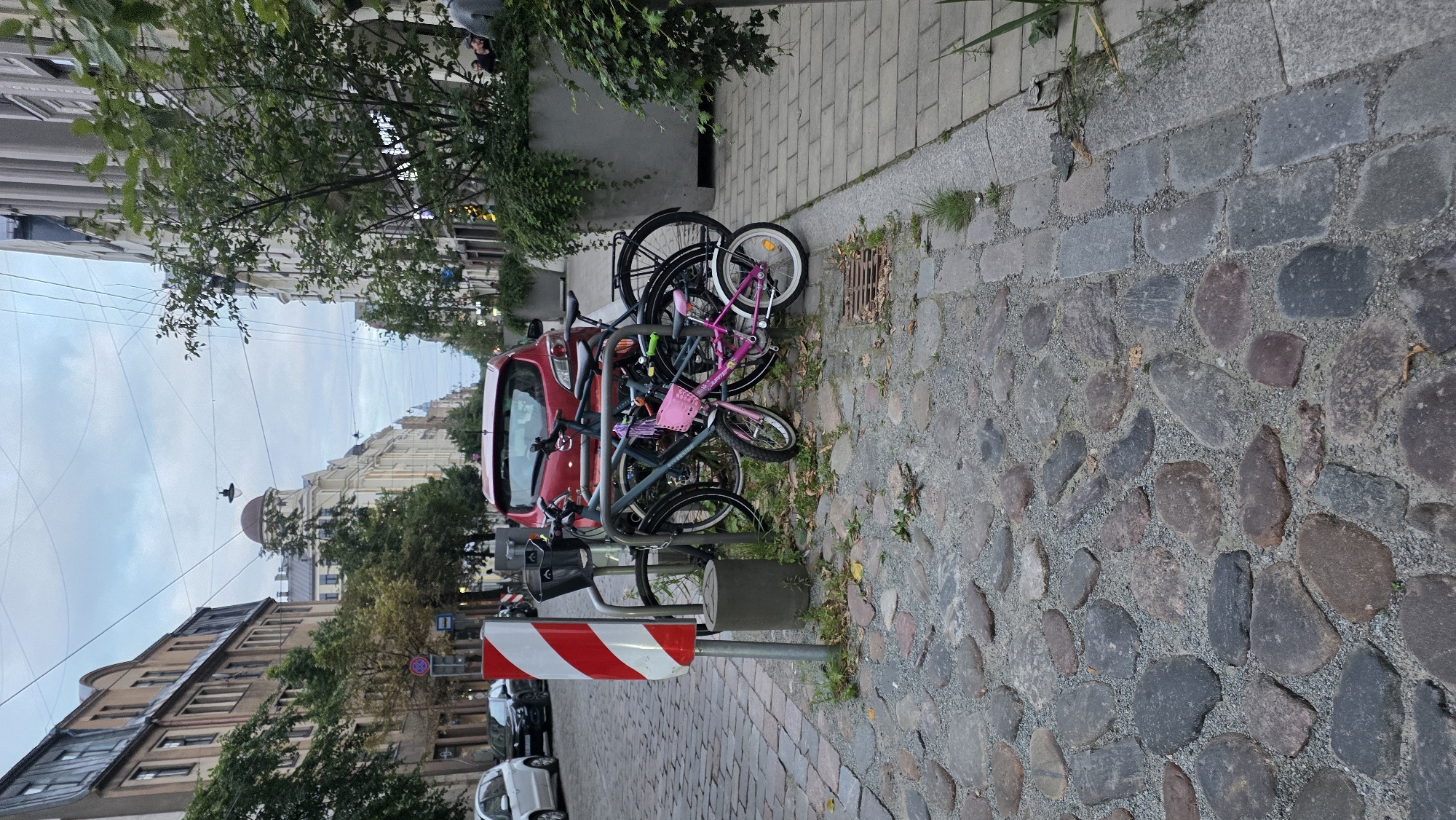 The image depicts a street scene in Riga, Latvia, on a cloudy day. In the foreground, a cobblestone street is visible, with a bike rack holding several bicycles. One bicycle is pink with a basket and streamers, and another is a darker color. A red car is parked behind the bicycles. A red and white striped traffic sign is mounted on a pole to the left of the bike rack. In the background, buildings line the street, some with arched windows and domed roofs. Green trees and planters are also present. The sky is overcast with a few power lines strung across it. There are no people clearly visible in the scene.
