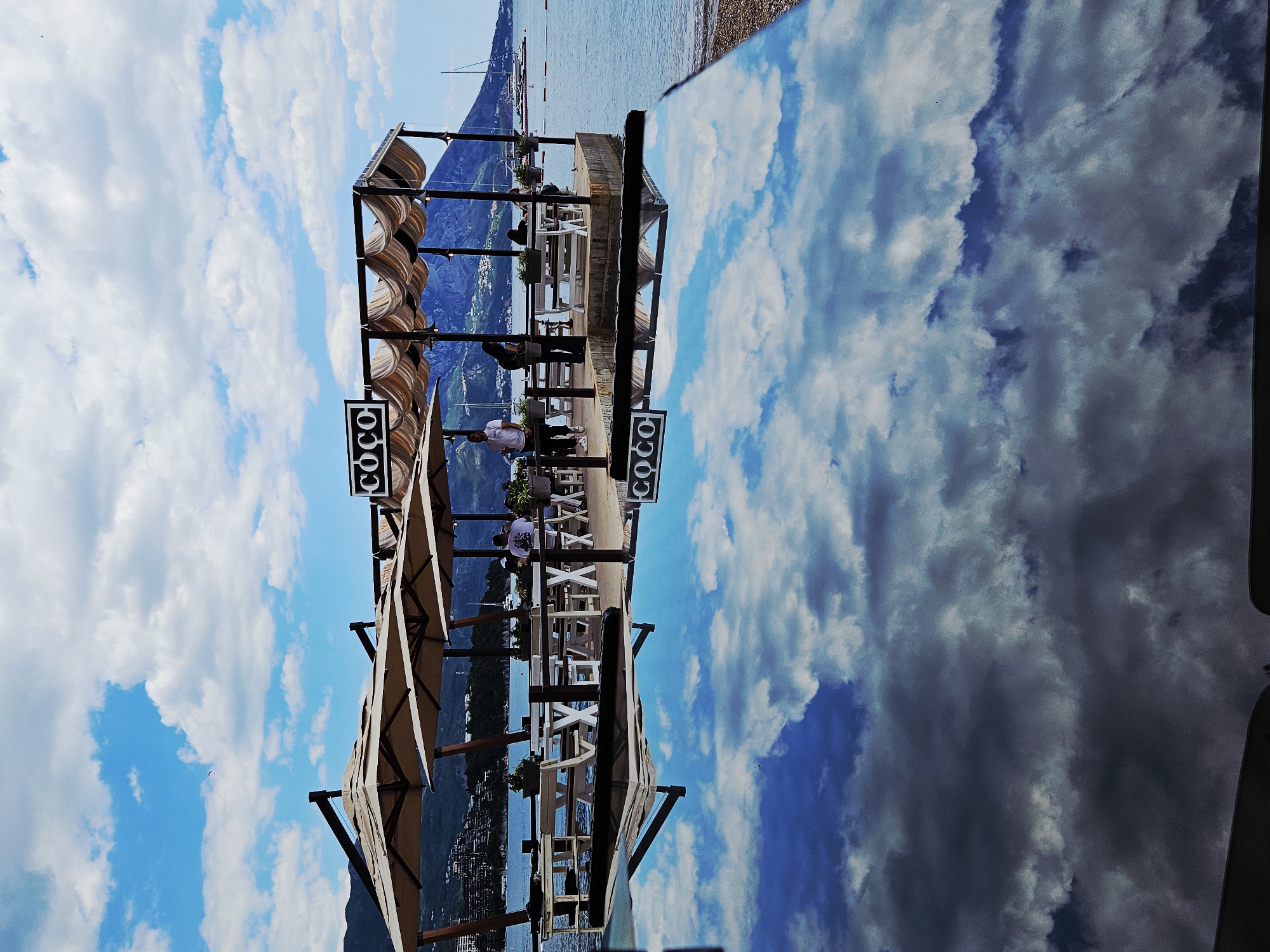 The image shows a scene at a beachfront location with a restaurant or bar named "Coco" featuring a covered seating area, set against a backdrop of mountains and the sea. People are seen sitting at the tables. To the right of the restaurant, there's what appears to be a large reflection of a cloudy sky, possibly on a reflective surface or billboard. The photograph appears to be taken in a sunny location.