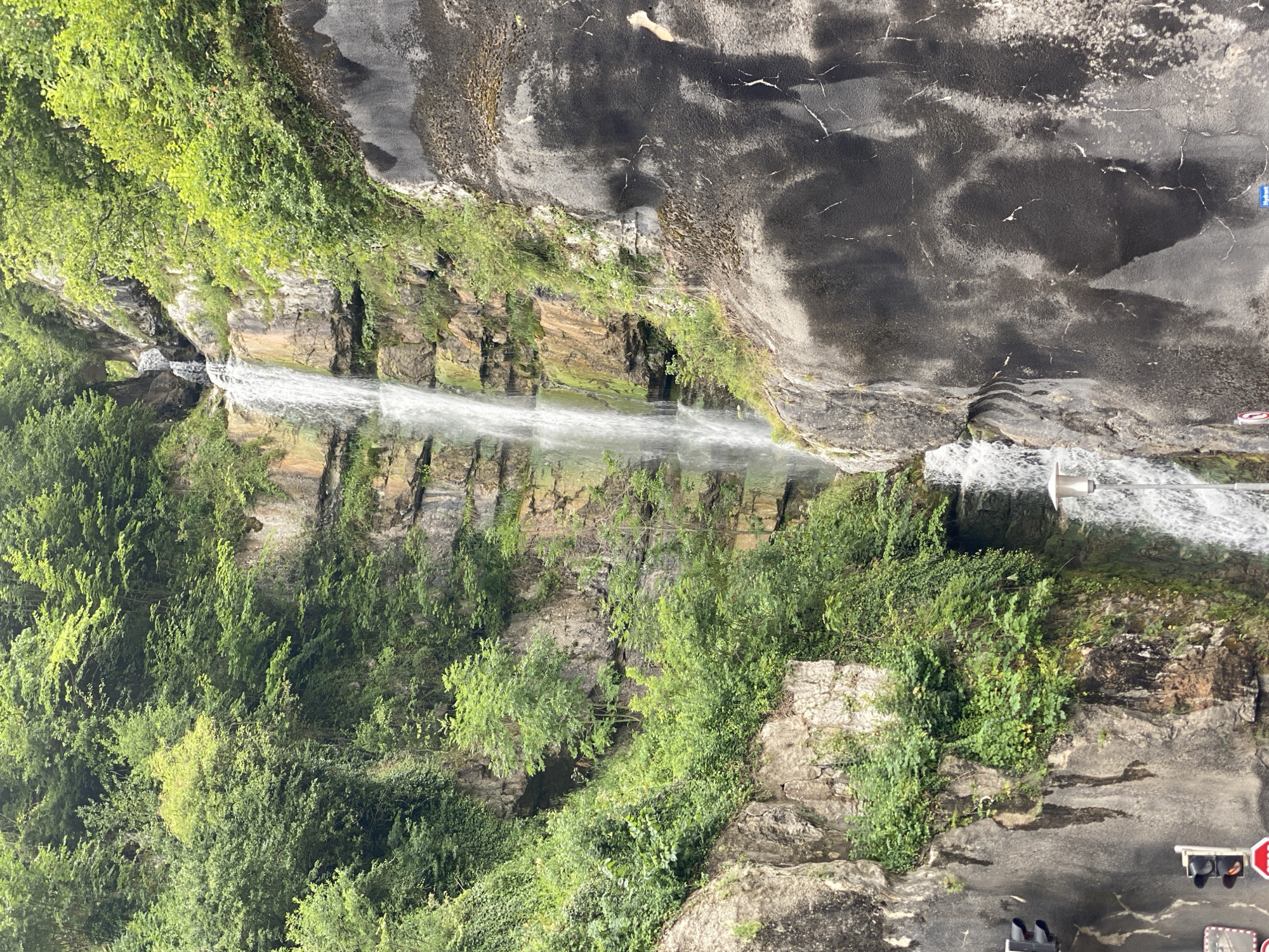 The image shows a tall waterfall cascading down a rocky cliff face covered in lush green vegetation. The waterfall is the central focus of the image, with water flowing in a narrow stream over various rock ledges. The surrounding cliffs are steep and uneven, with a mix of exposed rock and dense foliage. To the right of the waterfall, a dark, textured rock wall dominates the foreground, showing signs of weathering and possibly artificial reinforcement. In the lower left corner, traffic lights and a stop sign are visible, suggesting an urban or roadside setting. The overall environment appears to be a natural landscape integrated with man-made infrastructure. This waterfall is the "Märzenkamm Waterfall" in Hallstatt, Austria, a well-known natural landmark in the town.