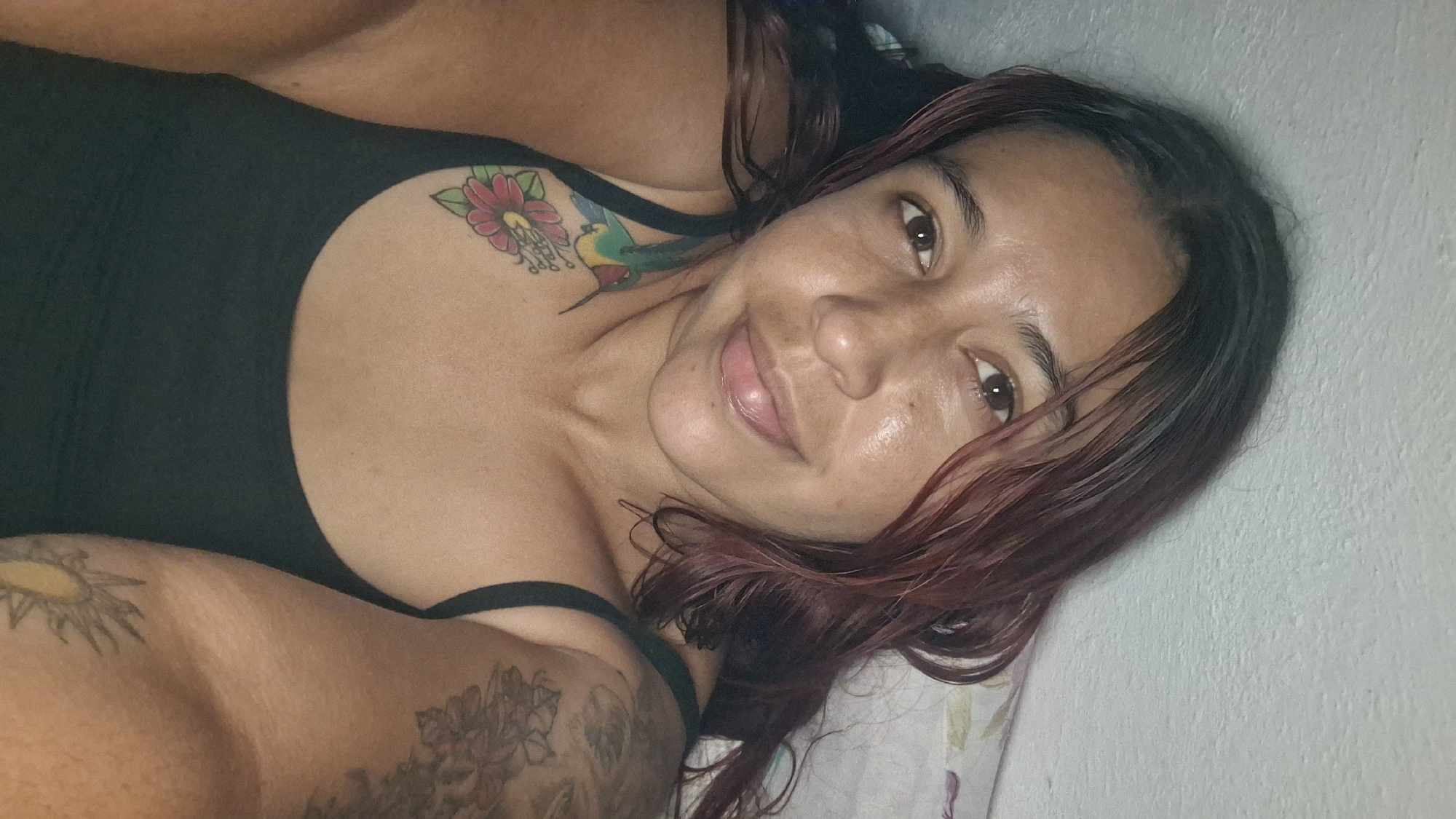 The image is a selfie of a woman lying down, looking towards the camera. She has dark hair with reddish highlights, and her eyes are brown. She is wearing a black tank top. There are tattoos visible on her left shoulder and upper chest, featuring a hummingbird and a flower. There is also a tattoo on her right arm. The background appears to be a wall and a bed with floral-patterned bedding. The lighting suggests an indoor setting, possibly at night or in a dimly lit room. There are no indications of the specific location within Vallecito, Venezuela.