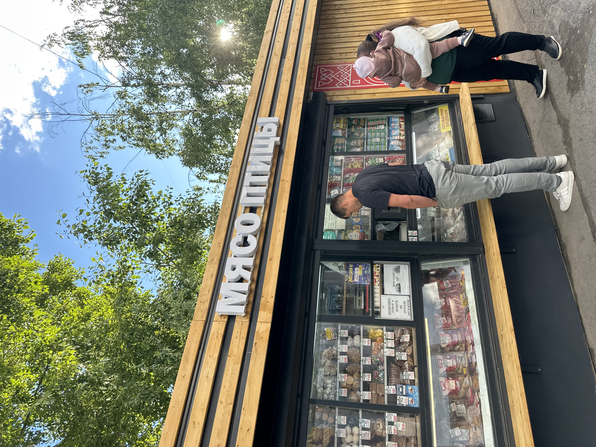 This image captures a street-level view of a small food shop with prominent Cyrillic signage that reads "МЯСО ПТИЦЫ" (Meat Poultry). The shop appears to be a butcher or deli, as the display windows are filled with various food items, including what looks like bread, sausages, and packaged meats, with price tags visible.
Two people are in the foreground. On the right, a person wearing a black t-shirt and grey pants is bending over, possibly looking at or interacting with the shop's display. On the left, a woman is carrying a child on her back. The child is wearing a light-colored jacket and a pink hat.
The shop itself has a modern facade with vertical wooden slats. Above the shop's entrance, there is a red sign with decorative white patterns. The overall scene suggests a sunny day, with dappled sunlight filtering through green trees that frame the top left of the image. The sky is blue with some white clouds. The ground in front of the shop appears to be asphalt or a similar paved surface.
Visible text on the shop windows includes price indications like "365" and "329-", and a sign that says "ПРОВЕРЯЙТЕ ТОВАР И ДЕНЬГИ, НЕ отходя ОТ КАССЫ" (Check goods and money without leaving the checkout). Another sign reads "АКЦИЯ" (Sale/Promotion) and "ОХЛАЖДЕННАЯ ПРОДУКЦИЯ" (Chilled Products).
The overall impression is that of a casual, everyday scene in an urban or suburban setting, likely during daytime.