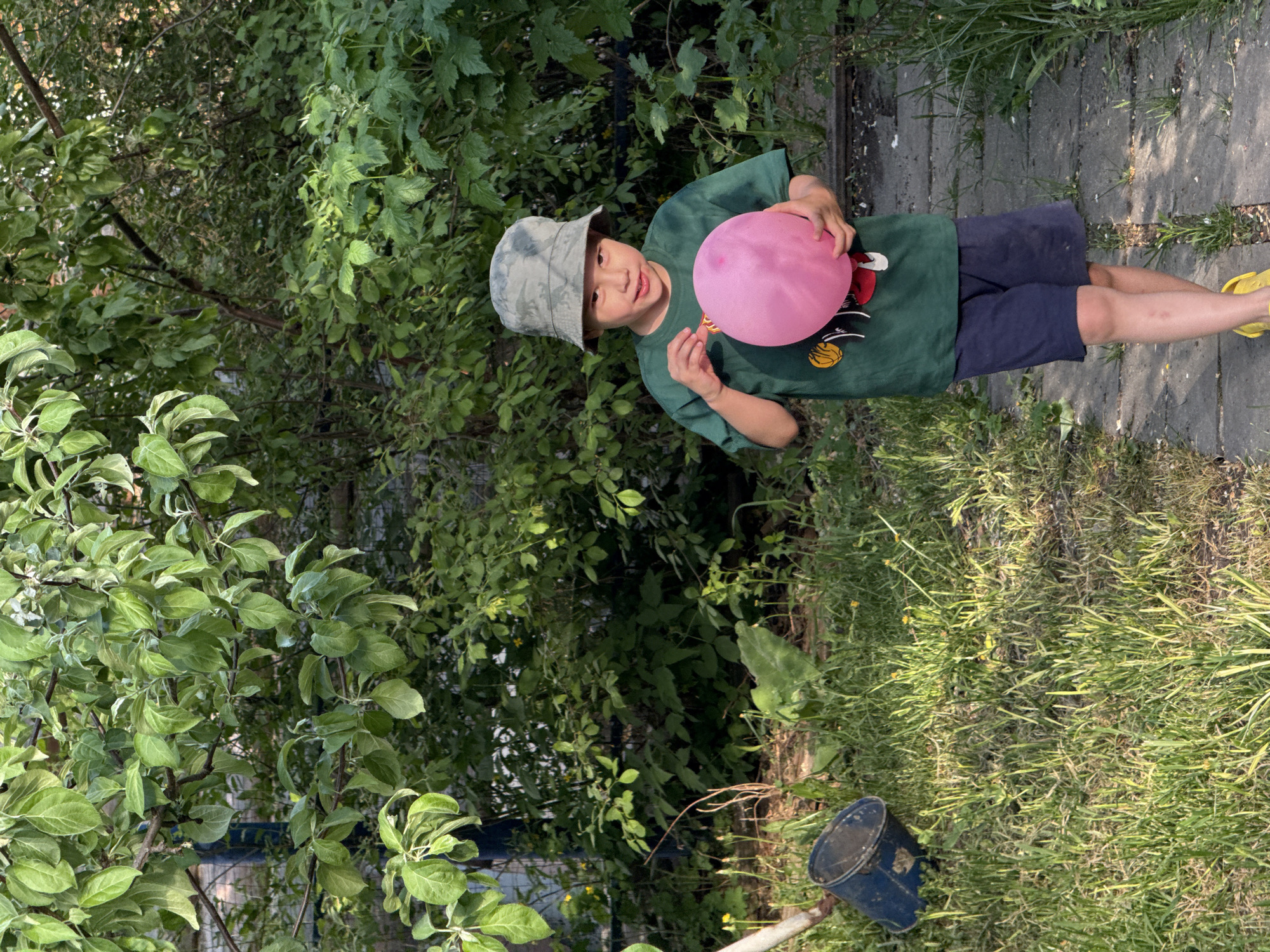 A child stands on a concrete path in a garden or yard, holding a pink balloon. He wears a green t-shirt with a graphic, dark blue shorts, and yellow sandals. A camouflage bucket hat covers his head. Lush green vegetation surrounds him, including trees, bushes, and grass. A blue bucket is visible in the lower right corner. The location is likely a residential area with ample greenery, potentially in the city of Chaikovsky, Russia, if that is the location where the picture was taken.