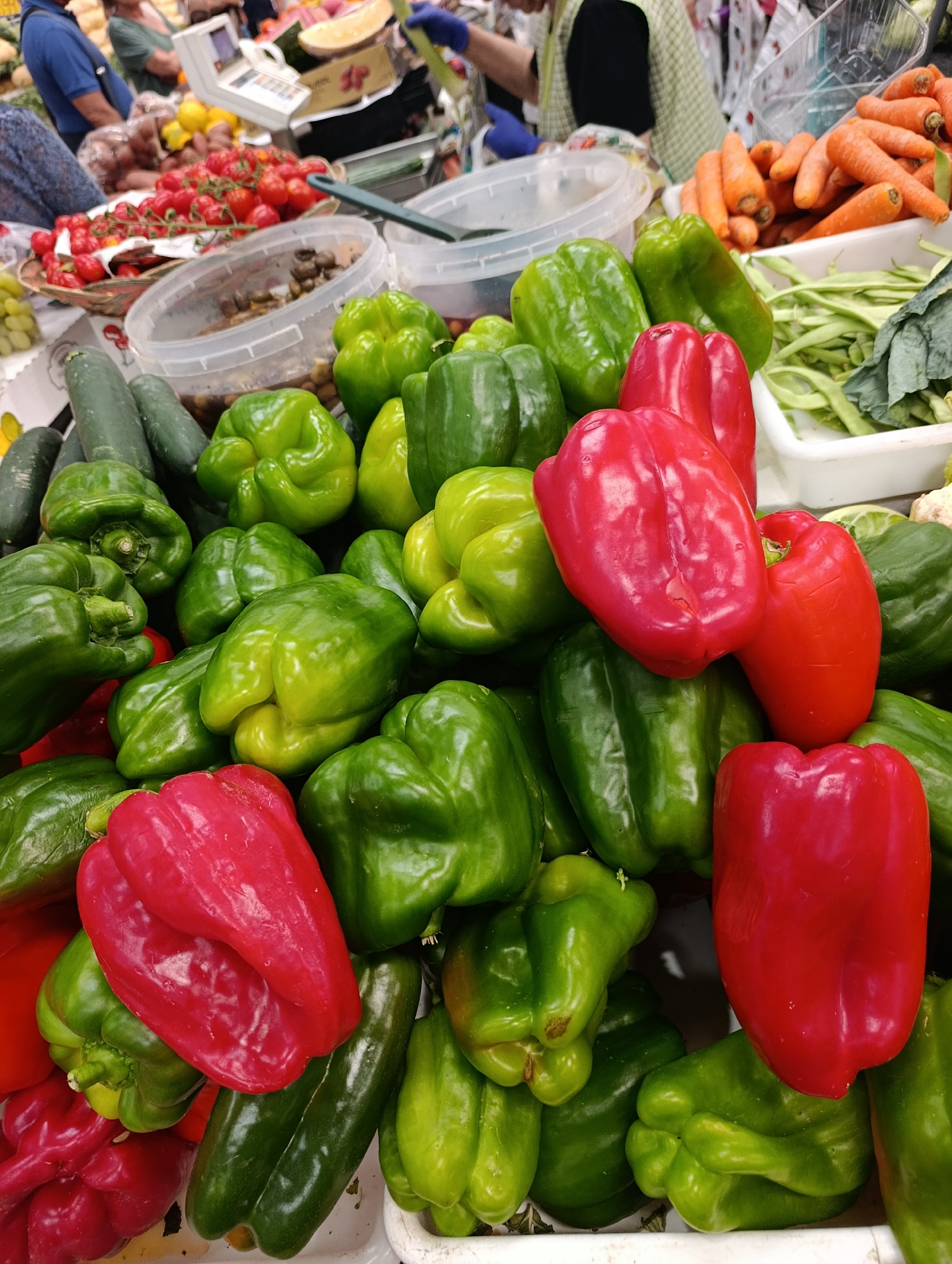 This is a close-up, eye-level, medium shot of a market stall in Setúbal, Portugal, overflowing with colorful bell peppers. The dominant subjects are a large assortment of green and red bell peppers, piled high in multiple layers and containers. Several cucumbers are visible in the upper left portion of the frame, also part of the produce display. In the background, out of focus, are hints of other market goods such as tomatoes and what appear to be carrots. The lighting is bright and natural, characteristic of an outdoor or well-lit indoor market. No people are clearly depicted or interacting with the produce, though the presence of a market stall implies human activity. The overall impression is of abundance and fresh produce at a local market.