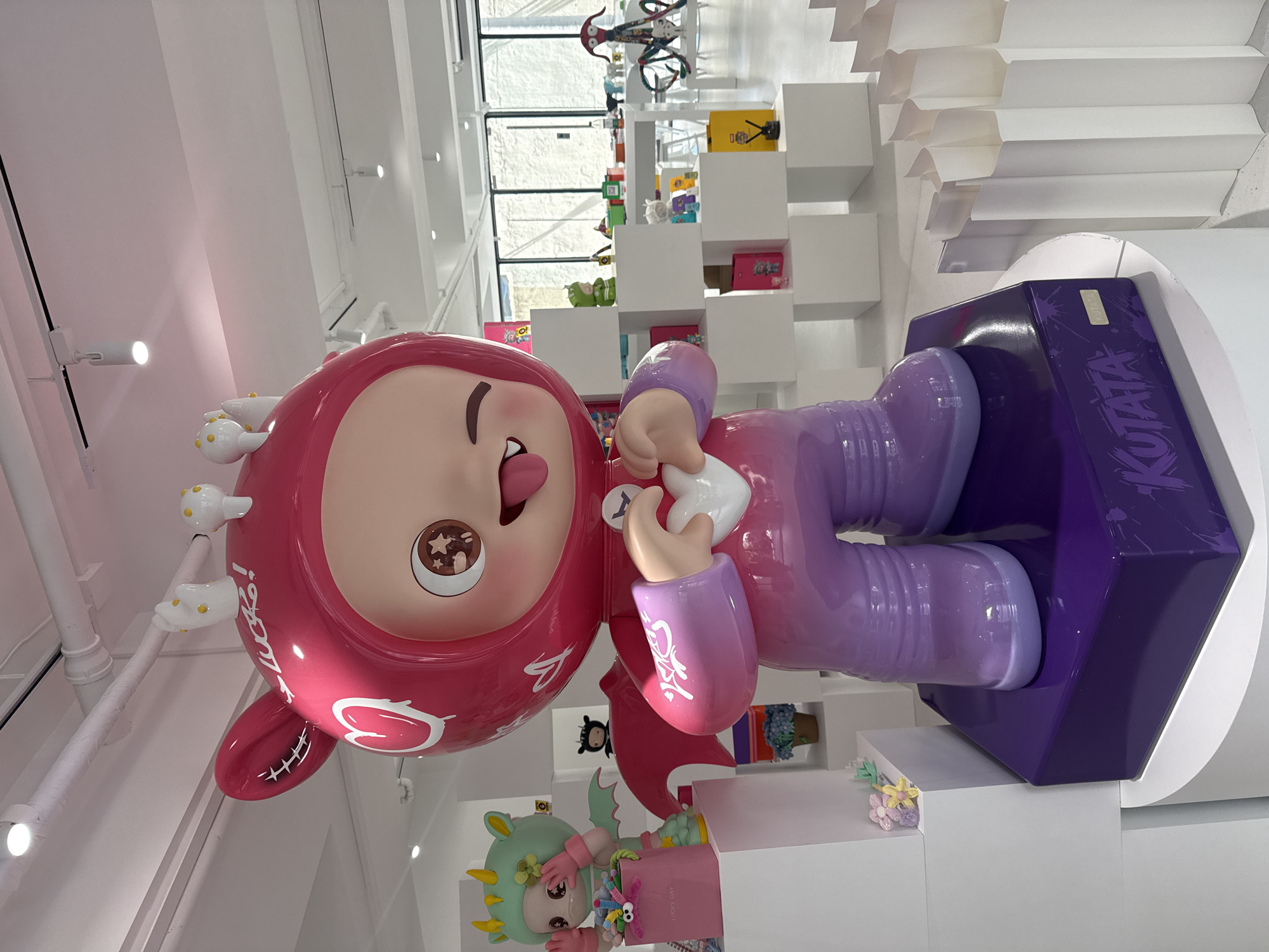 The image depicts a large, vibrant pink and purple cartoon character statue standing on a purple base labeled "KUTATA" in a bright, modern retail store. The statue has a hood with bunny ears, a large expressive face with one eye winking and tongue sticking out, and is forming a heart shape with its hands. The character appears to be a collectible toy. The store environment features white shelving units displaying smaller figurines, colorful boxes, and decor. In the background, large windows offer a view of an outdoor area with a bicycle. The store is located in Shanghai, China.