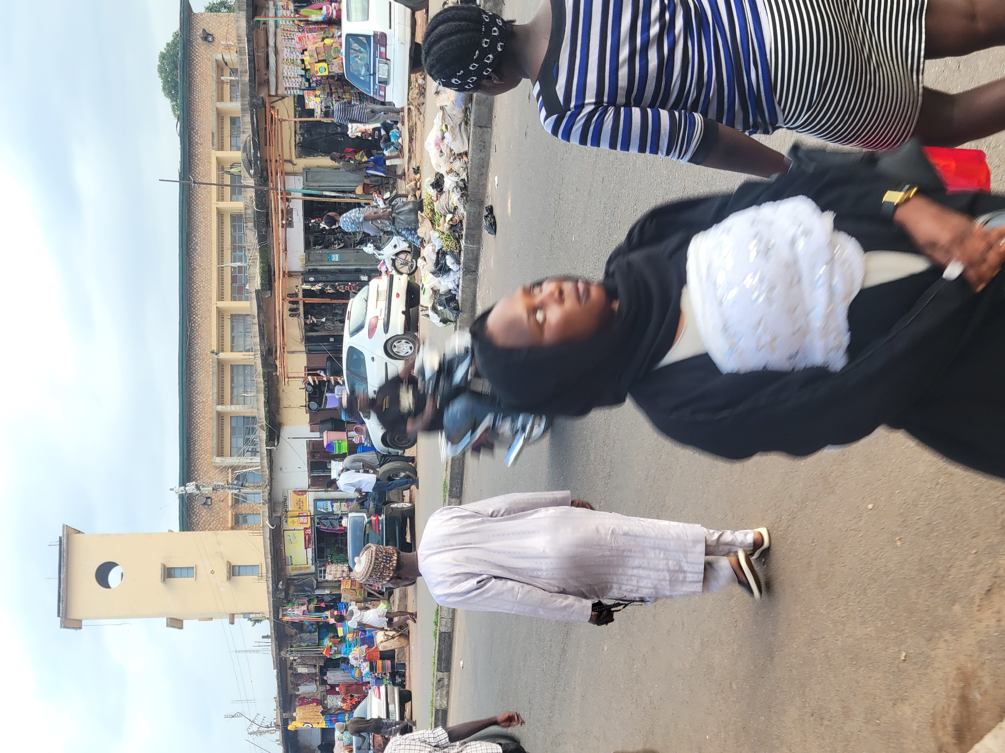 The image depicts a bustling street scene in Jos, Nigeria, likely in a market area. Several people are visible, including a woman in a black and white striped shirt and a black headscarf, and another person in a light-colored abaya and cap. A woman in the foreground is wearing a black hijab with a white, embellished fabric piece draped over her. The street is lined with shops and stalls displaying a variety of goods, including what appear to be food items, clothing, and household products. Several vehicles are also present, including a white car and motorcycles. The ground is paved, and there is a significant amount of refuse piled along the side of the street, suggesting an informal waste management situation. The buildings appear to be multi-story structures, some with balconies. One prominent building features a tall, light-colored tower with a circular opening. The sky is overcast, suggesting it might be a cloudy day. The overall impression is one of lively commercial activity and everyday life in a Nigerian urban setting.