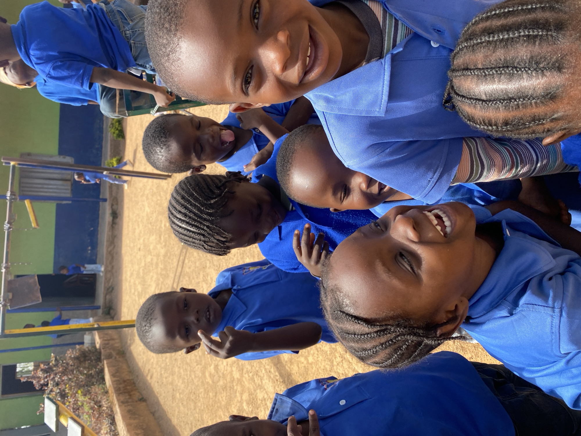 A group of children wearing blue shirts and dark pants are outdoors in Gura Top, Nigeria. They are gathered closely together, looking up at the camera. Some are smiling, laughing, or sticking out their tongues. The scene is bright with sunlight. In the background, there is a playground with equipment and a building painted green and blue. The ground is sandy.