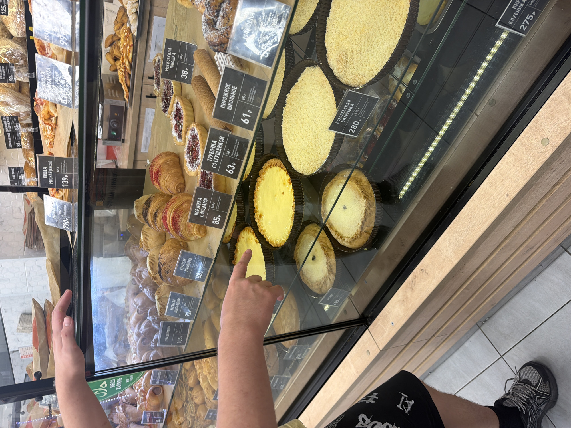 A person's hand points towards a display case filled with baked goods in a store. The display case contains various pastries, including croissants, tarts, and cakes. Several price tags are visible in front of the baked goods, some of which appear to be in Russian. In the background, shelves are stocked with more bread and baked items. The overall environment suggests a bakery or a grocery store with a bakery section. The presence of Russian text on the price tags suggests the location is likely in Russia.