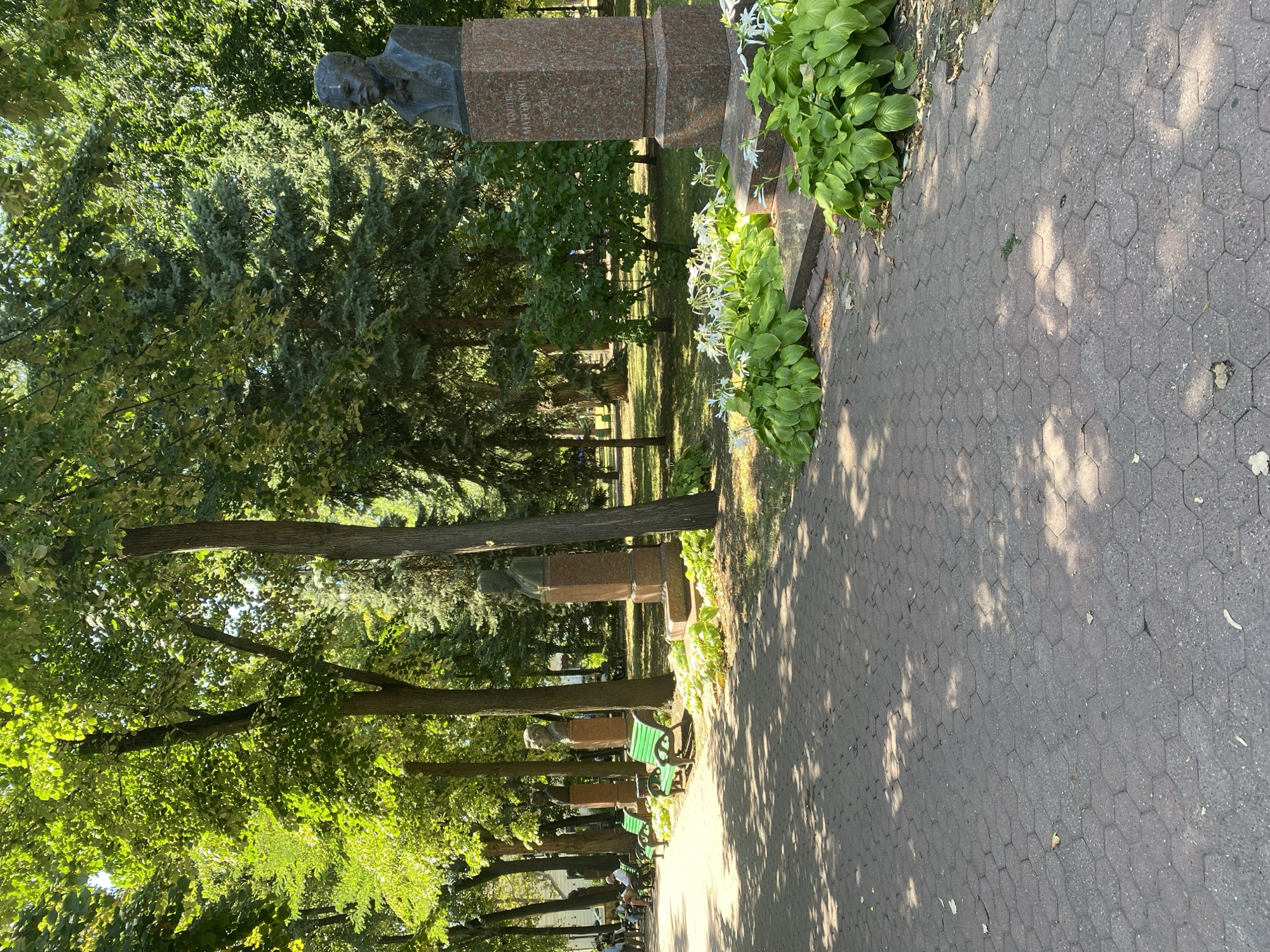 The image shows a memorial in Chișinău, Moldova, likely located in a park or public square. The scene features a paved walkway with hexagonal bricks leading towards a line of busts on pedestals. To the right, a prominent bust of a man with a suit and tie is visible, situated on a tall, red granite pedestal with an inscription. In front of this bust, there are lush green plants and white flowers. Further down the walkway, more busts are visible, each on similar pedestals, interspersed with green park benches. A row of mature trees with dense green foliage forms a backdrop, casting dappled shadows on the path. In the distance, on the left side of the image, a few people are seated on benches, enjoying the outdoors. The overall environment suggests a well-maintained public space with a focus on historical commemoration and greenery.