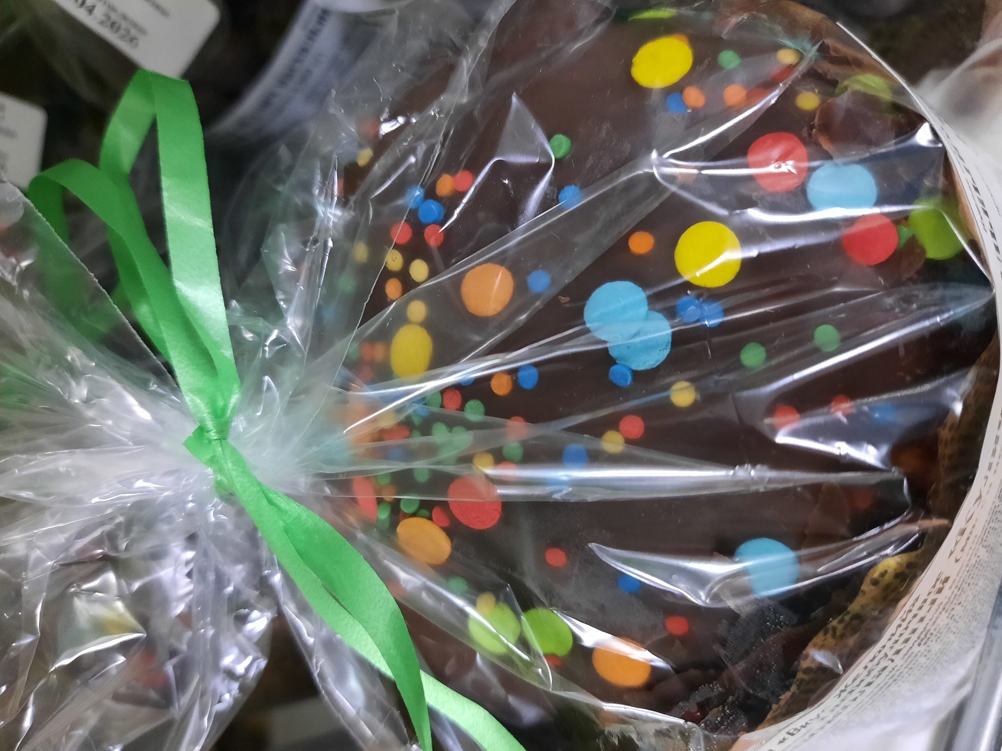 The image shows a chocolate lollipop in a clear plastic wrapper tied with a green ribbon. The lollipop is decorated with colorful candy dots. Part of Krasnogorskoye, Russia can be seen through the clear wrapper.
