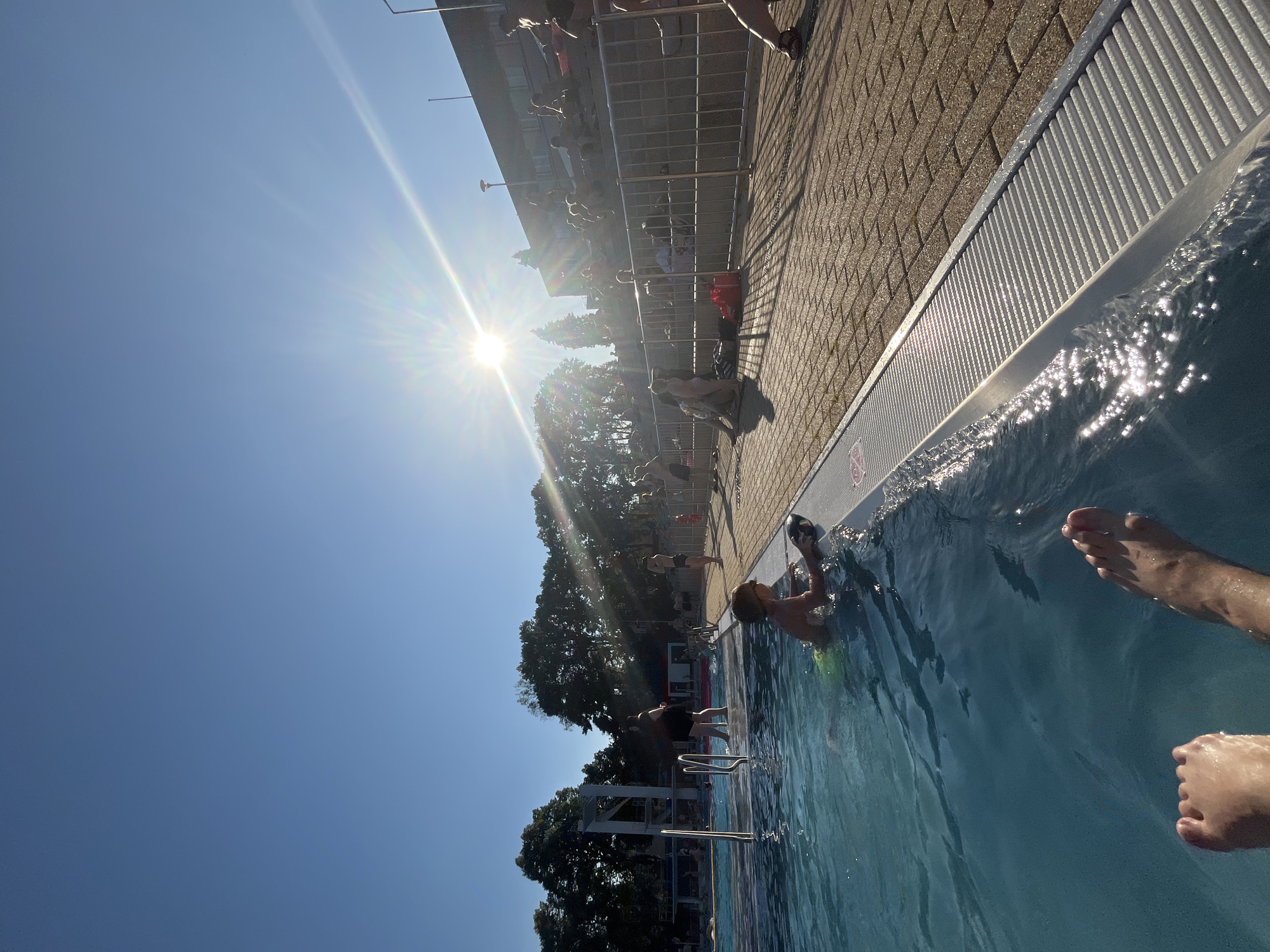 The image depicts a sunny day at an outdoor swimming pool. The sun is bright and high in the sky, casting lens flares and illuminating the water, suggesting it is midday. The pool area is lined with brick paving and a metal grate drain system along the edge. Several people are visible in and around the pool. Some are in the water, with their legs and arms visible, while others are standing or sitting by the pool. One person is partially submerged in the water, reaching for the edge. The atmosphere appears relaxed and recreational, with people enjoying a summer day. Trees are visible in the background, suggesting an outdoor setting. A building structure with railings is also present behind the trees. The image is captured from a low angle, looking upwards towards the sun and the pool. There is no visible text in the image.