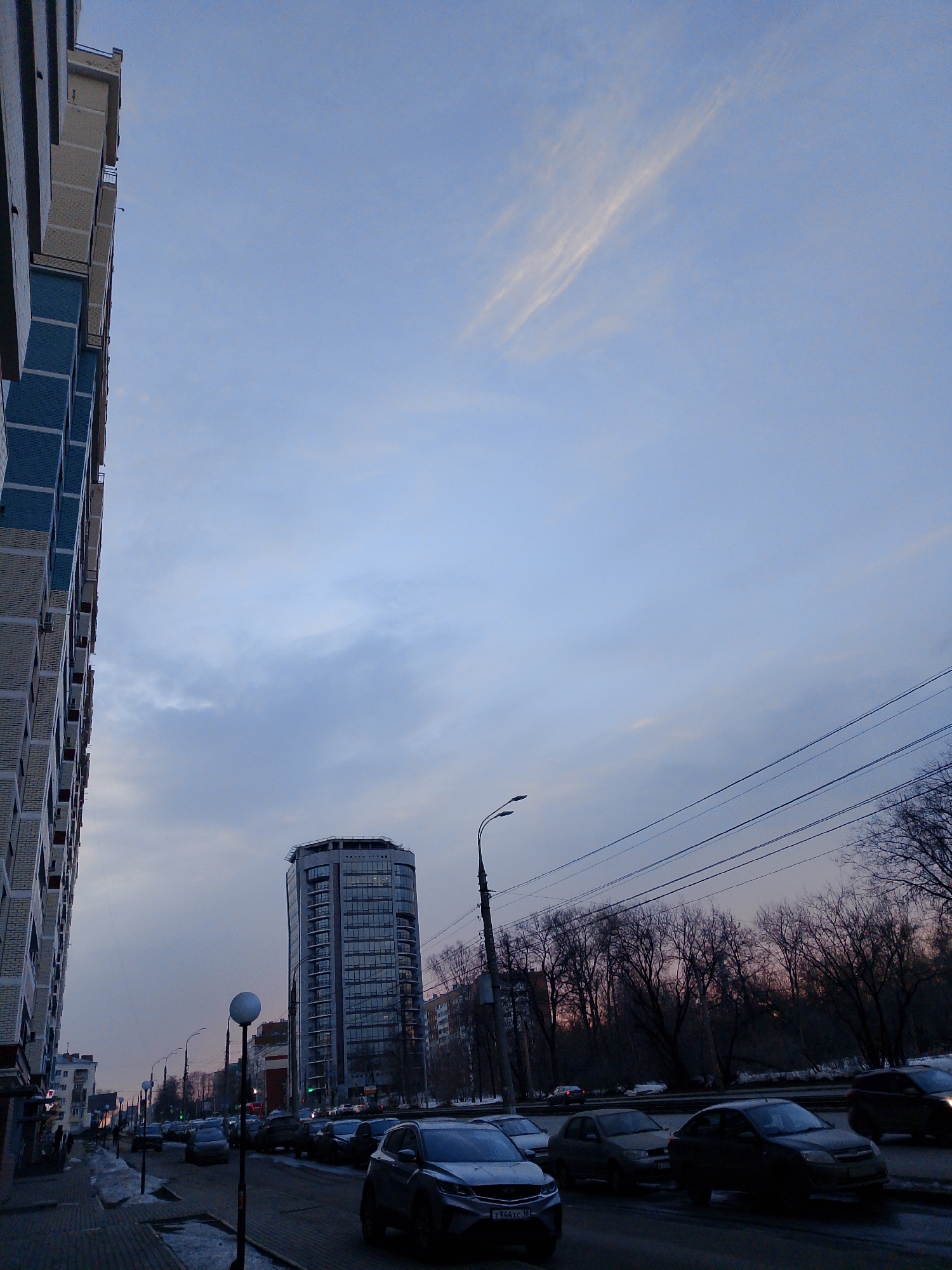 The image depicts a street scene in Izhevsk, Russia, at dusk. A tall, modern glass-and-steel building stands prominently in the midground, while a multi-story residential building is partially visible on the left. The street is lined with parked cars. Bare trees suggest it is winter or early spring. Utility poles and wires are visible overhead. The sky is a mix of blue and grey, indicating the time of day. Small piles of snow are present on the sidewalk, and the pavement appears wet. There are no clearly visible people in the image.