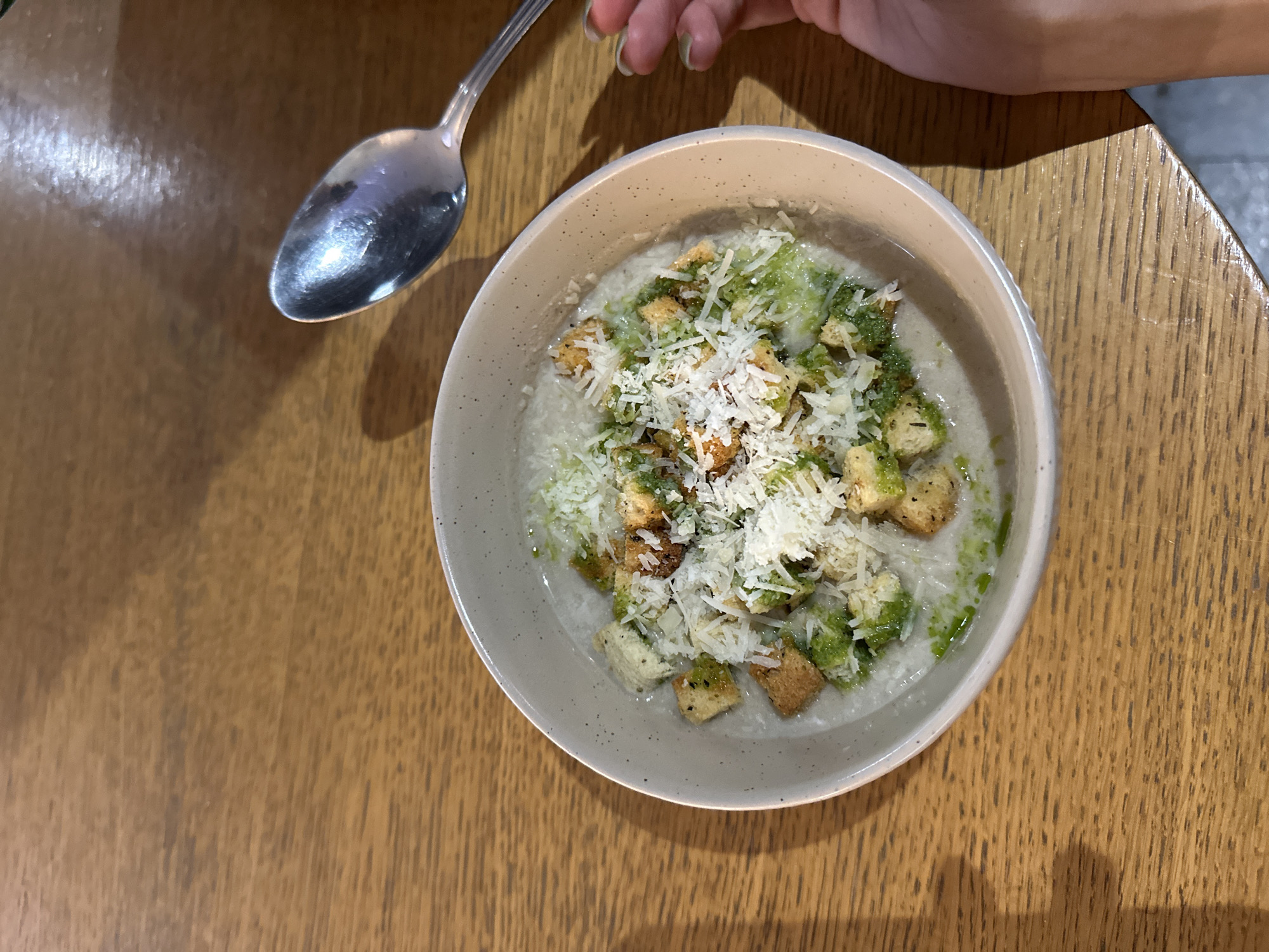 The image presents a top-down view of a meal served on a wooden table, likely indoors, in Novi Sad, Serbia. The primary subject is a light-colored bowl filled with food. The dish appears to be a creamy soup, risotto, or porridge, with a light base. It is generously topped with several ingredients: a vibrant green, oily substance, possibly pesto or a herb oil, is drizzled over the surface. Scattered across the dish are numerous golden-brown croutons, adding texture. A substantial amount of white, grated cheese, resembling Parmesan, covers the croutons and green topping.
To the upper left of the bowl, a silver spoon with a reflective surface is resting on the table. In the upper right corner, a person's hand is visible, holding the handle of the spoon. The hand has slender fingers, and one fingernail visible has light green nail polish. The hand is positioned as if about to eat, or having just taken a bite. The wooden table beneath the bowl and spoon features a clear grain pattern, with some reflections visible on its polished surface, particularly near the spoon. The lighting suggests an indoor setting, with some shadows cast by the hand and spoon. No text is visible in the image.