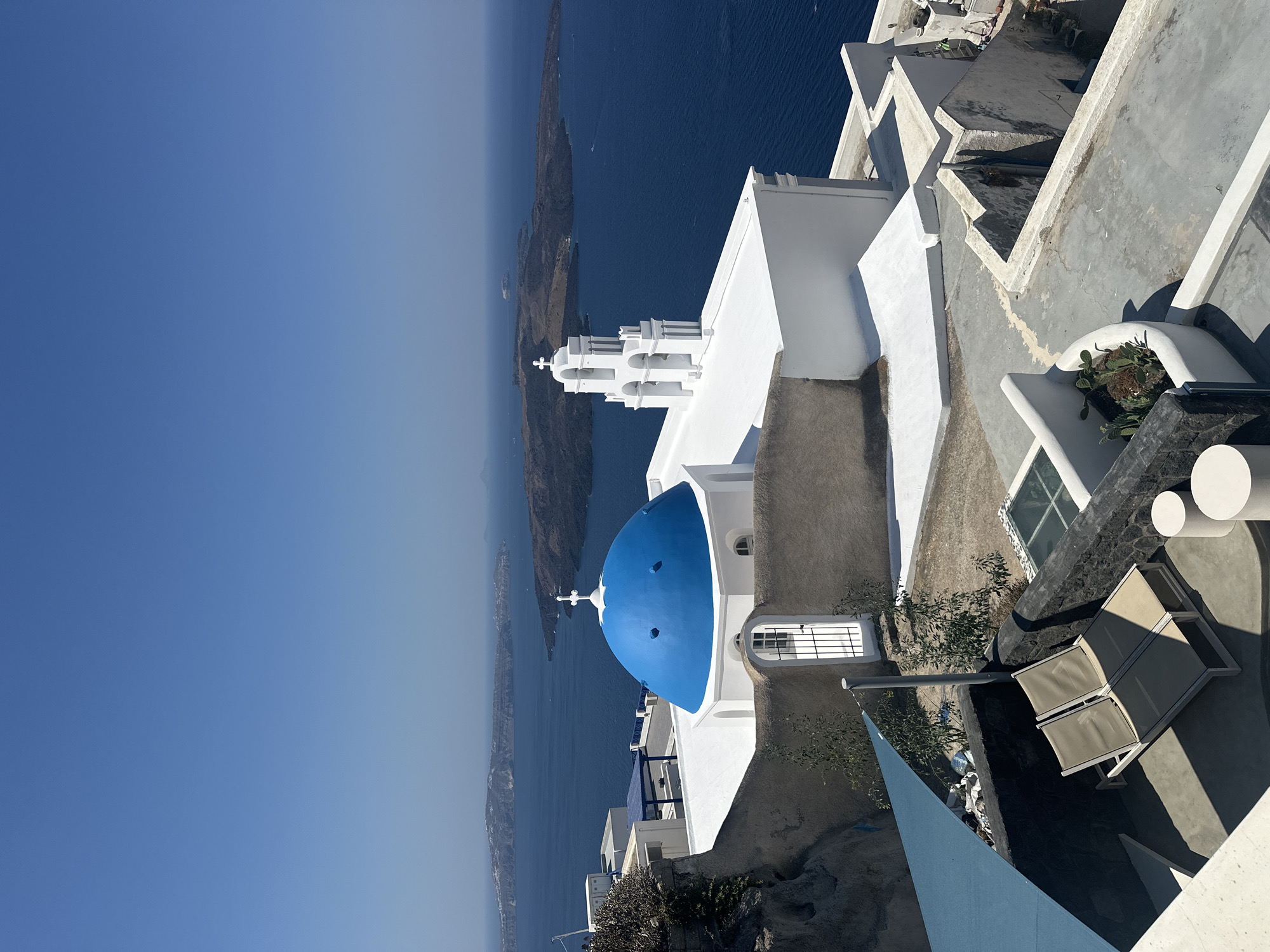 This is a high-angle, full shot of the Aegean Sea and a white building with a bright blue dome, located in Oia, Santorini, Greece. The sky is a clear, bright blue, with no clouds. In the foreground, there are lounge chairs on a patio. The building features a prominent blue dome and a white bell tower with arched openings. The sea is a deep blue and is dotted with several islands in the background. The architecture is characteristic of the Cycladic islands, with whitewashed buildings and blue accents. The lighting suggests a bright, sunny day.