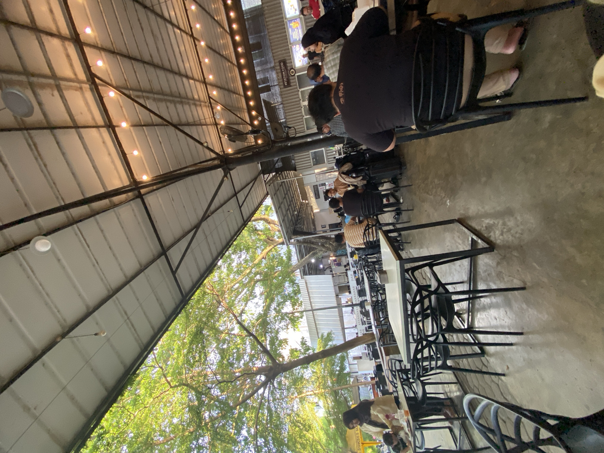 The image shows an outdoor dining area, possibly in Lhokseumawe, Indonesia, with people seated at tables under a large, open-air metal roof structure. The roof is supported by dark metal beams and has string lights hanging from it. A large tree with green foliage is visible in the background, adding a natural element to the scene. People are engaged in eating and socializing. Rows of tables and chairs extend into the distance. The ground appears to be concrete. There are overhead fans attached to the roof supports.