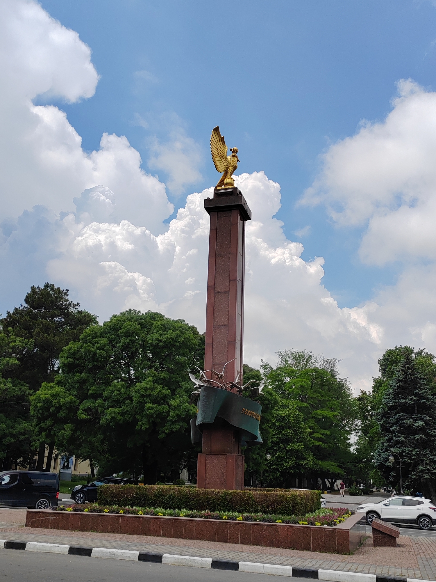A monument featuring a golden bird sculpture atop a tall, reddish-brown column stands in a park-like setting with lush green trees. The monument has a decorative base with a sculpted ribbon and bird figures. In the foreground, a sidewalk and street are visible, with black and white painted curbs. A black van and a black car are parked on the side of the street. In the background, people are walking in the distance, and a white car is driving on the road. The sky above is blue with large, fluffy white clouds.