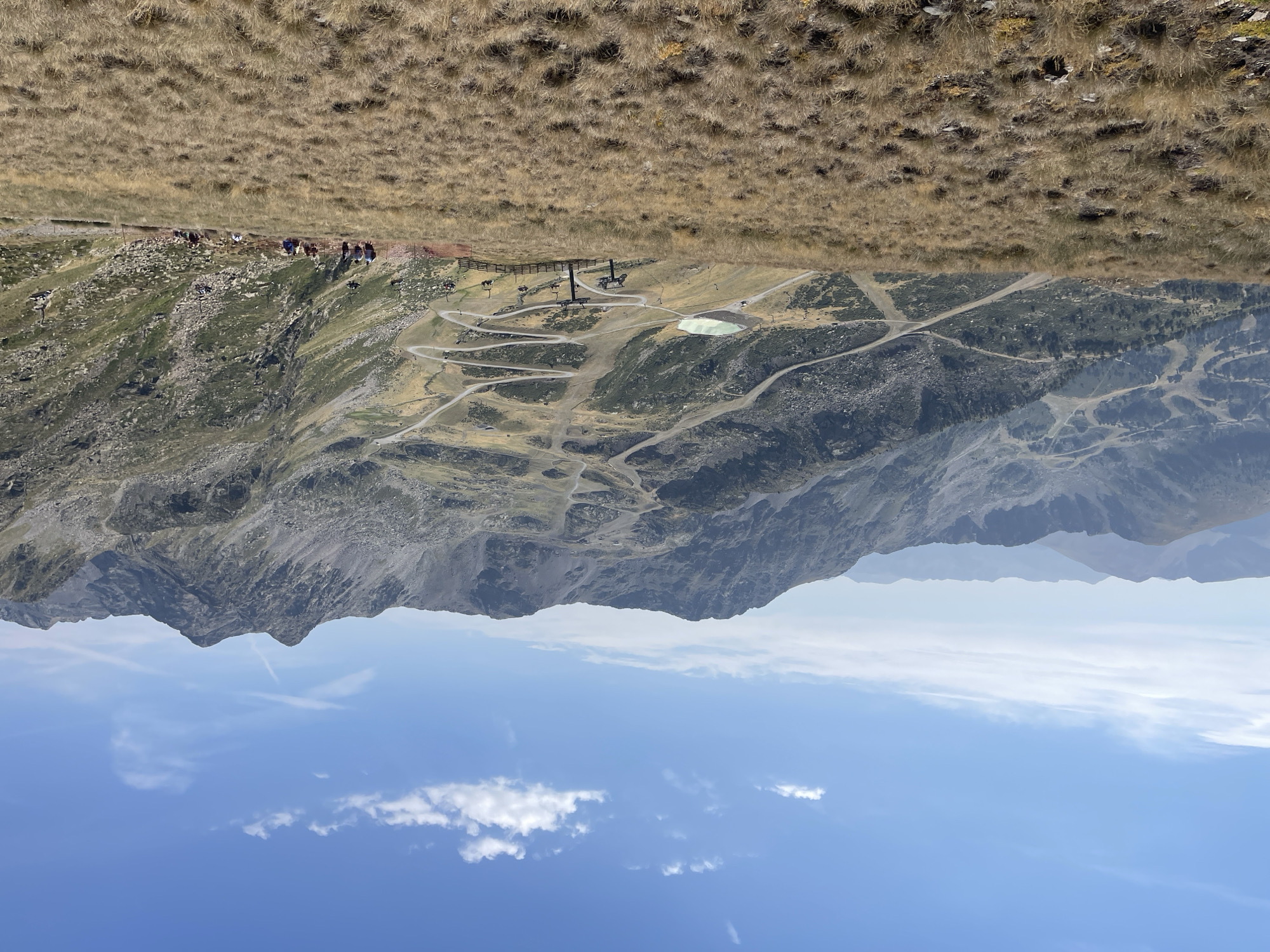 The image shows a wide, mountainous landscape under a clear blue sky with scattered white clouds. In the foreground, dry, golden-brown grass covers a sloping hill. In the midground, a winding road or ski slope snakes down the mountainside, with several chairlifts visible. A small, turquoise-colored body of water is situated near the road. A group of people, appearing as small figures from this distance, are walking on the grassy slope in the midground. The background consists of rugged, rocky mountains stretching into the distance, with more winding paths and vegetation visible on their slopes. The overall scene depicts a mountainous region, possibly a ski resort in the summer or off-season, with a focus on the natural landscape and recreational infrastructure.